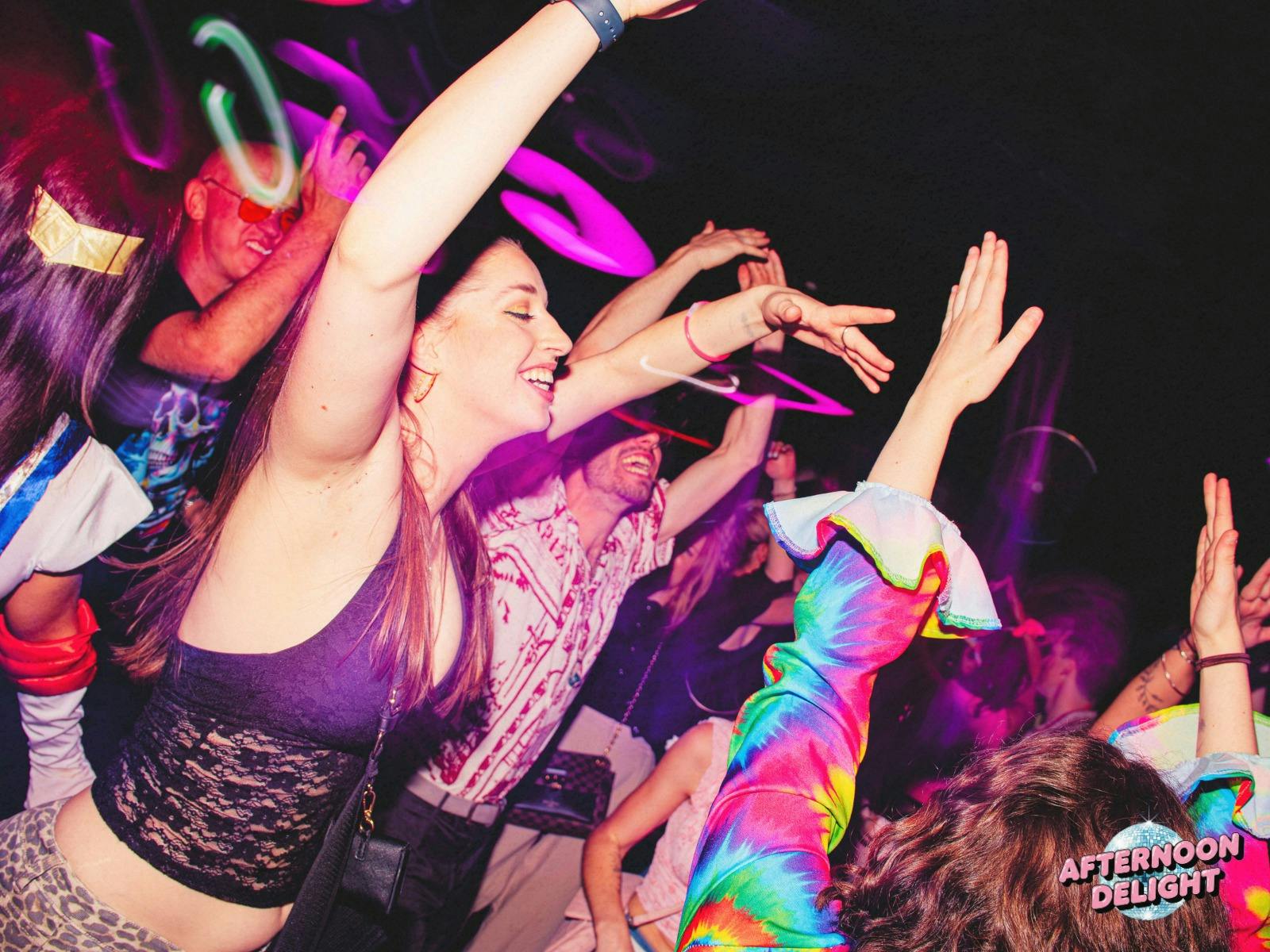 Image shows colourful energetic dancefloor crowd with thier hands in the air, neon lights in back