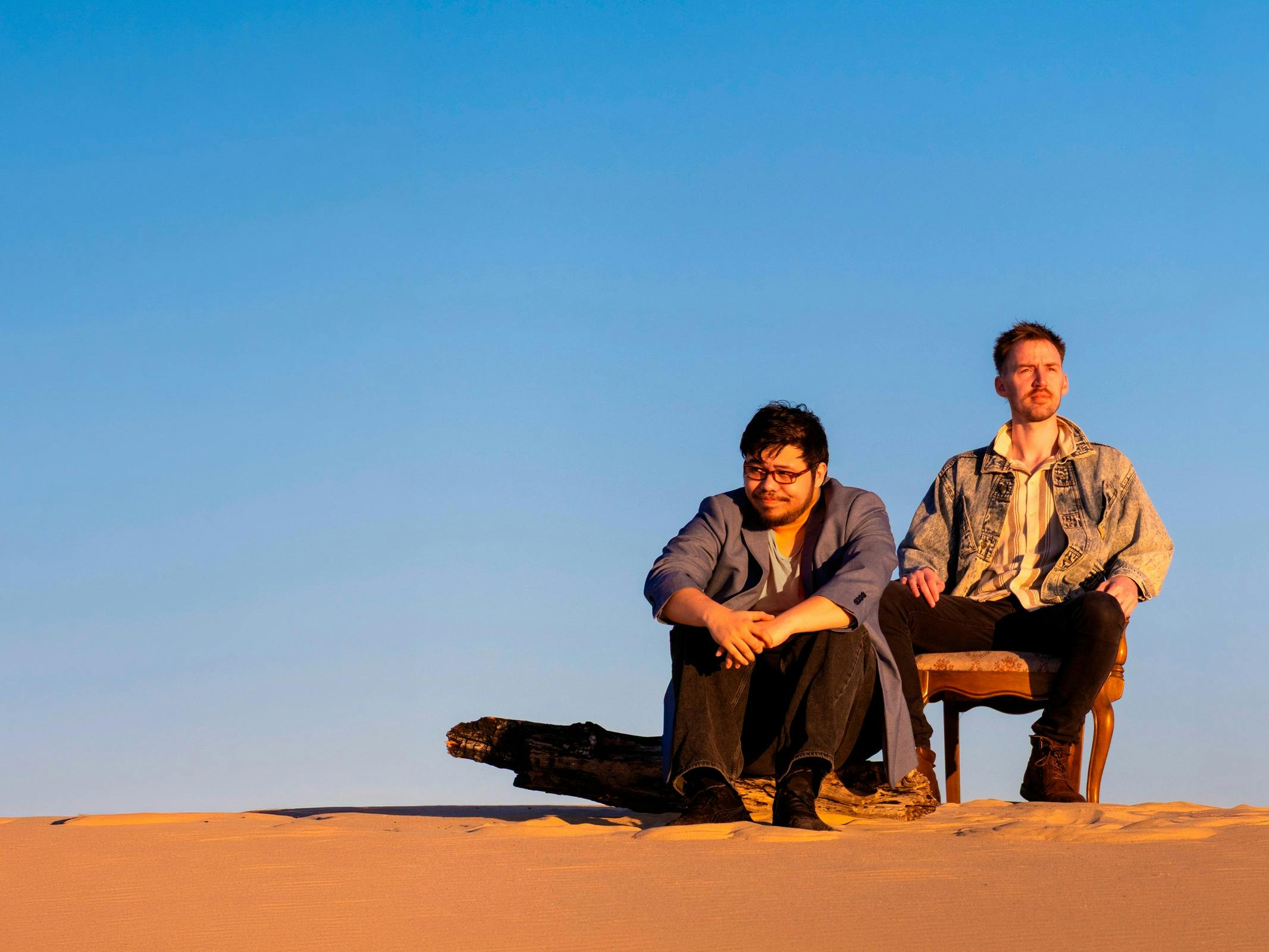 Two men seated in a desert landscape beneath a blue sky.