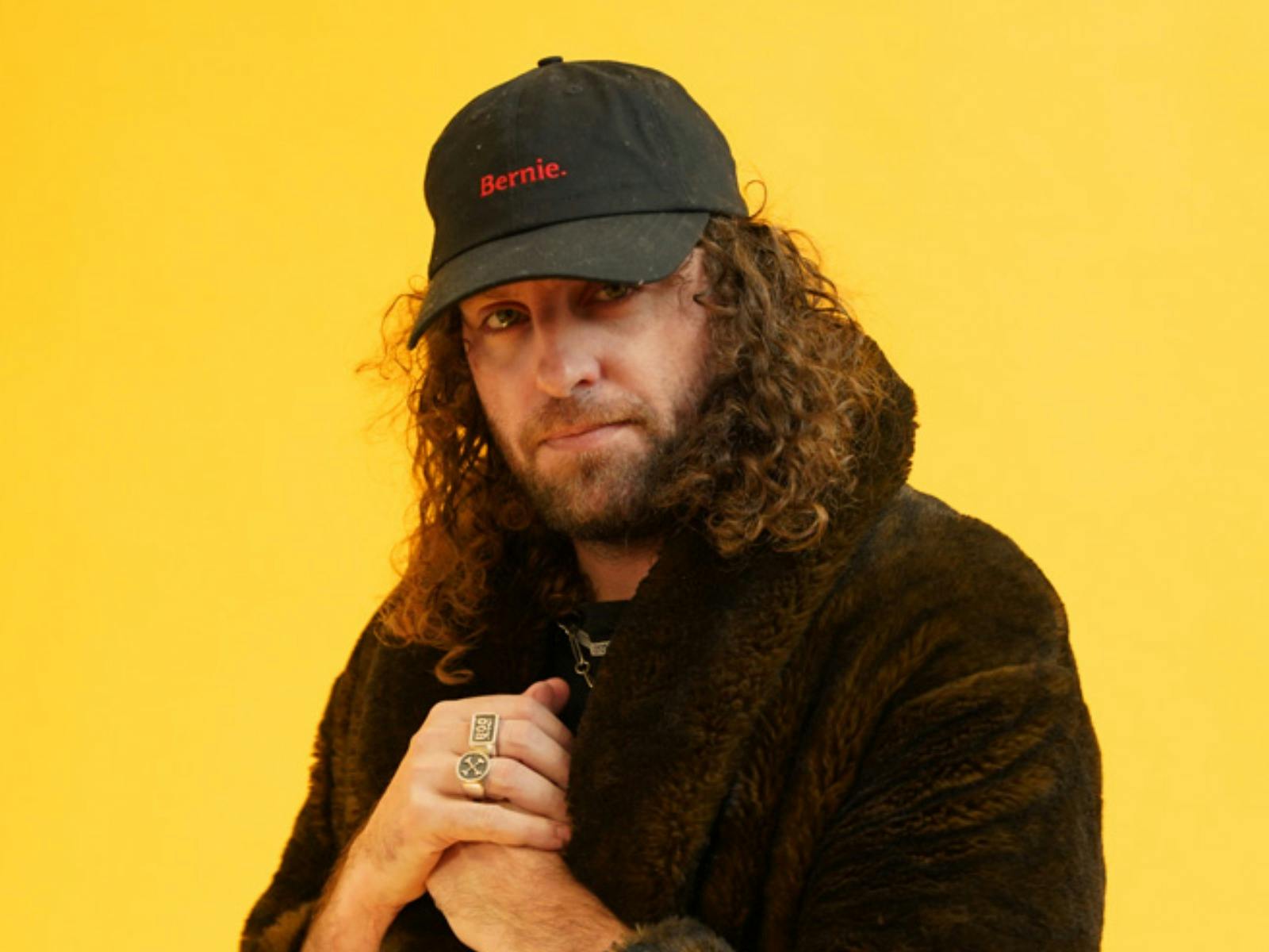 man with brown shoulder length hair and black cap on with yellow background holding his shirt collar