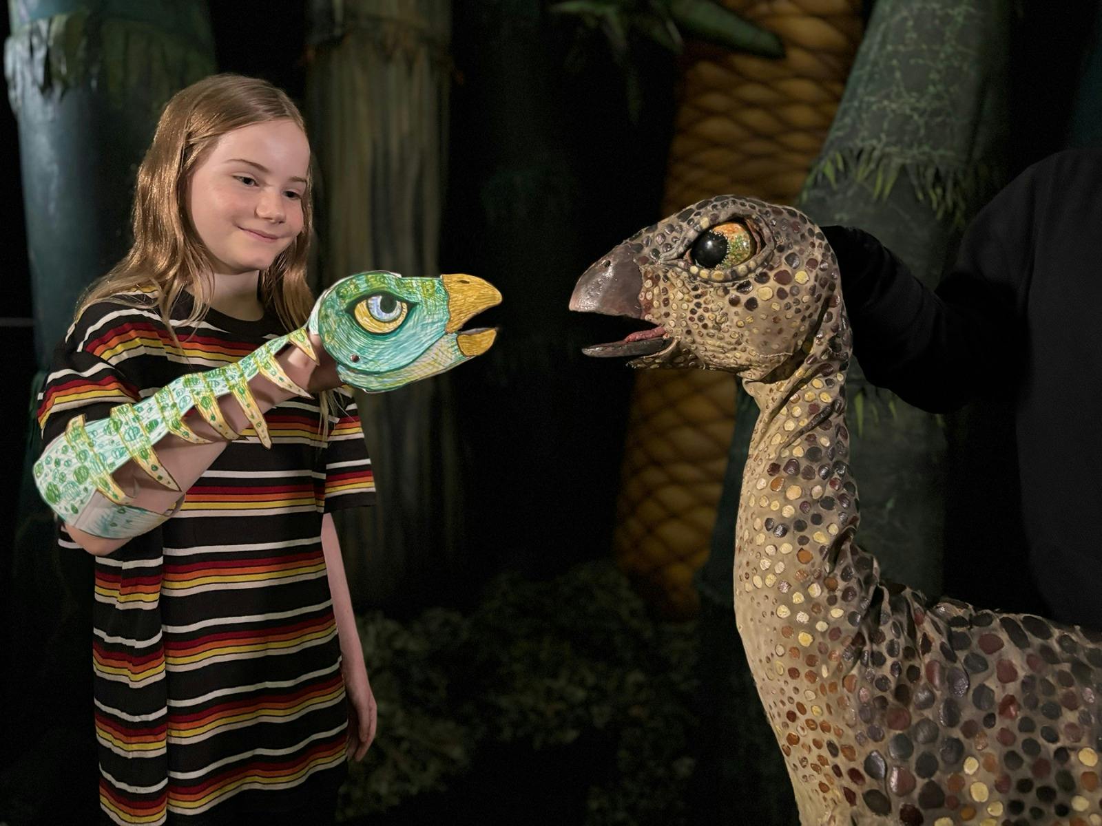 A girl wearing a striped T-shirt plays with a paper dinosaur puppet while facing a dinosaur puppet