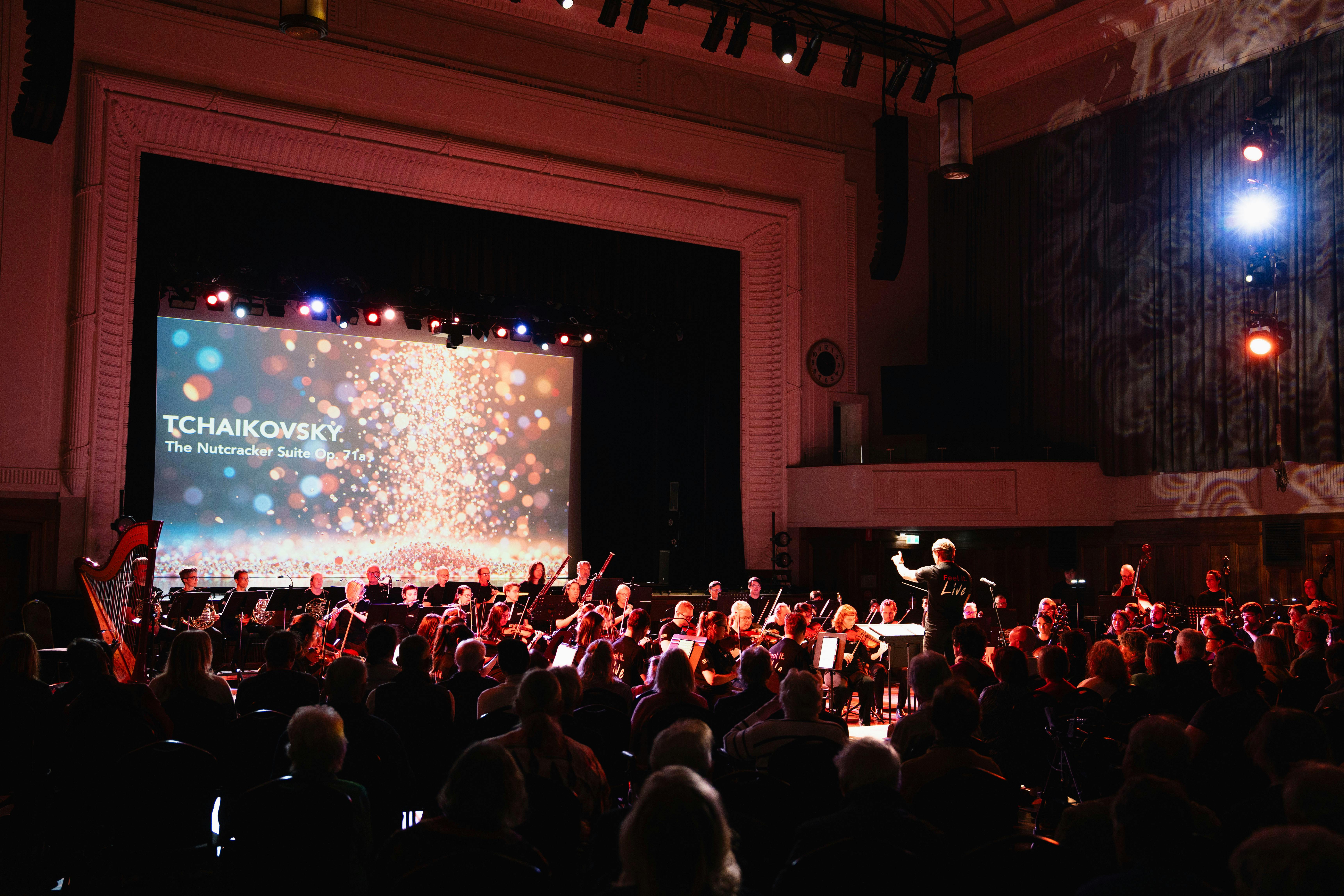 Orchestra performing on stage in front of an audience in a hall, bathed in a warm red light