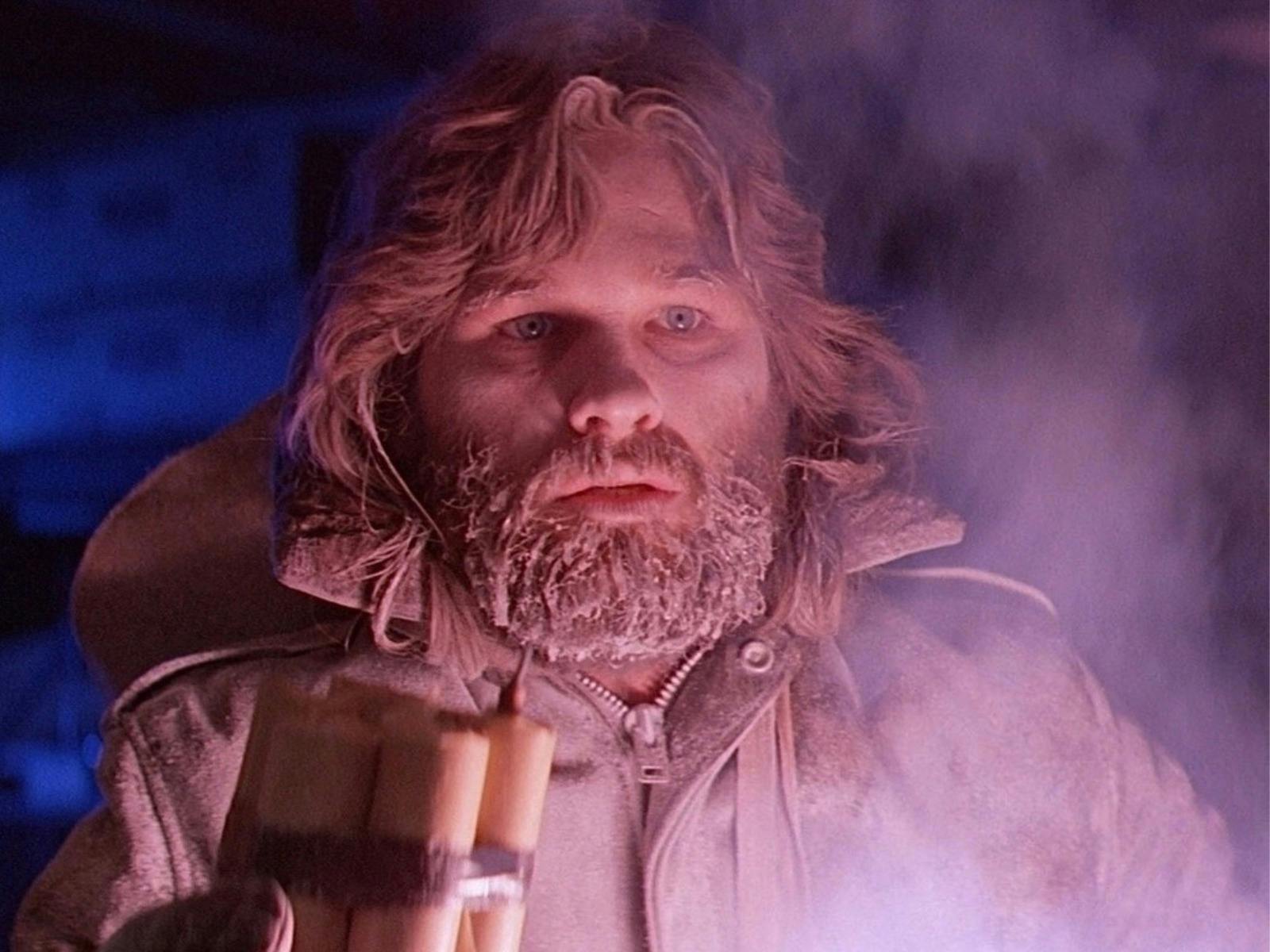 A man in a snow jacket stands with an icy beard looking just right of the camera, lit by flame