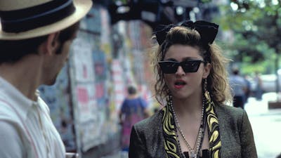 Woman walking towards man wearing 1980s style clothing, sunnies and big hair bow