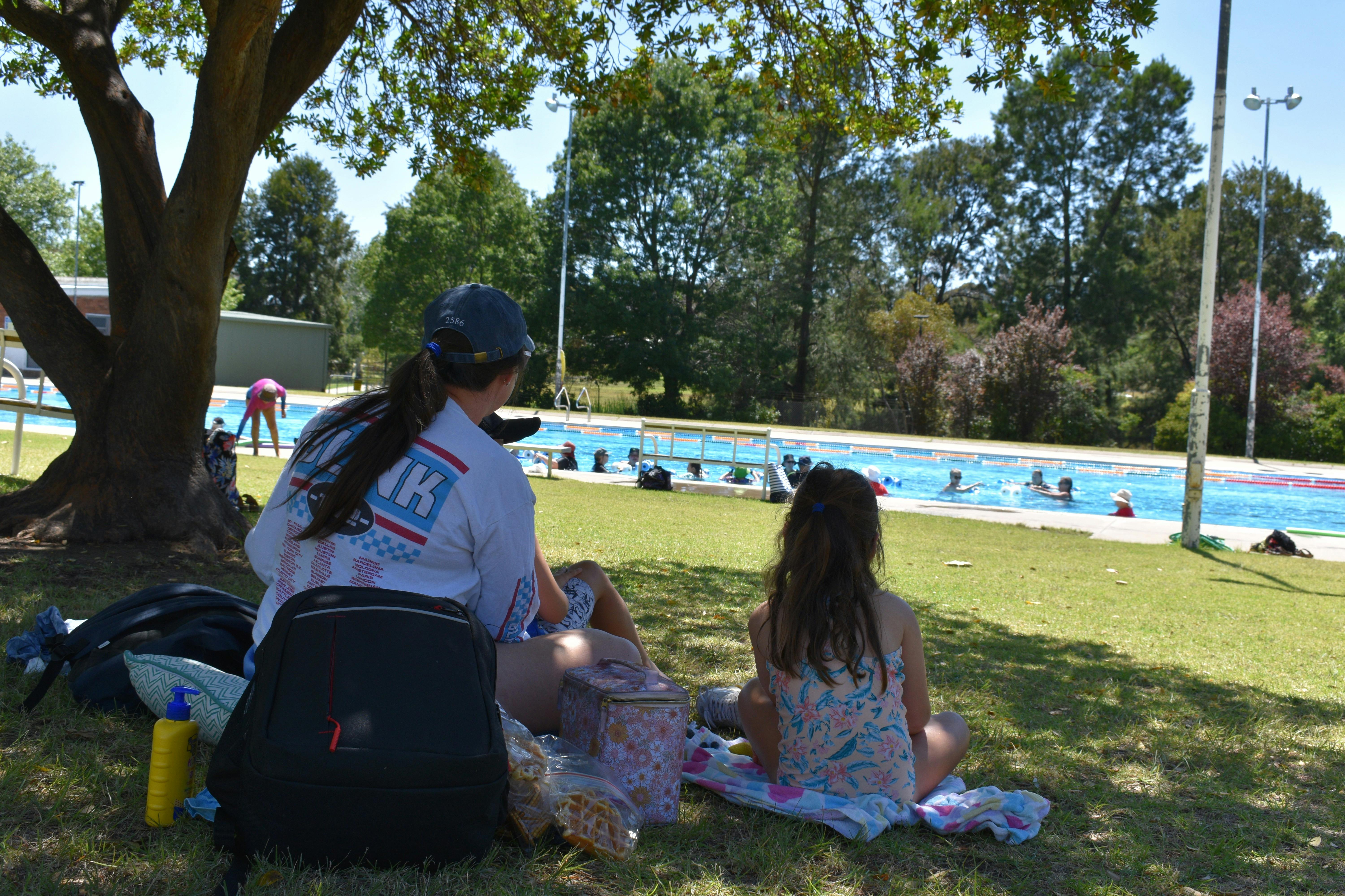Mother and child resting in the shade of a tree, looking out at the busy pool.