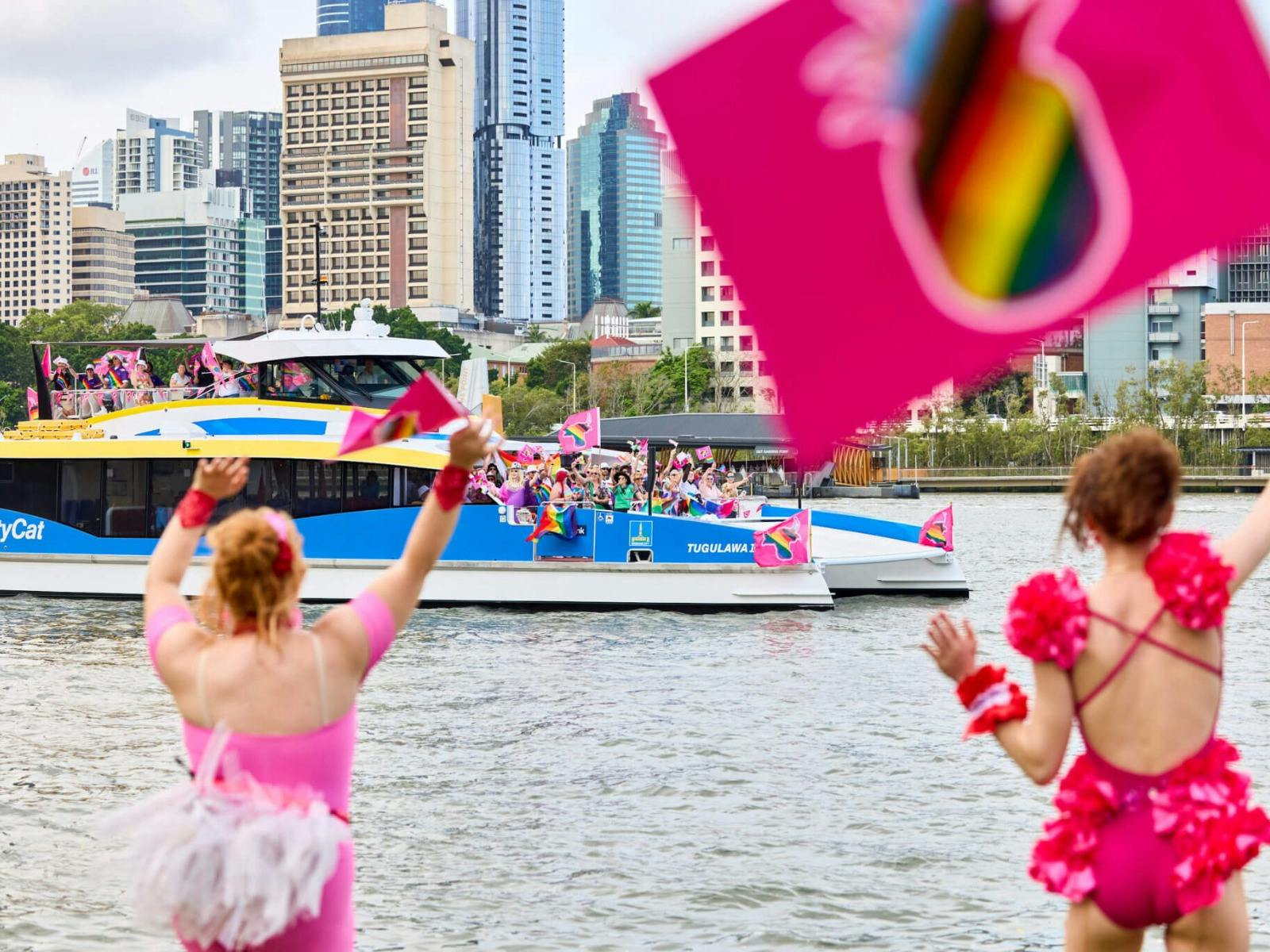 Two people in vibrant pink outfits cheer as a CityCat with colorful flags passes by on the river. Two people in vibrant pink outfits cheer as a CityCat with colorful flags passes by on the river.