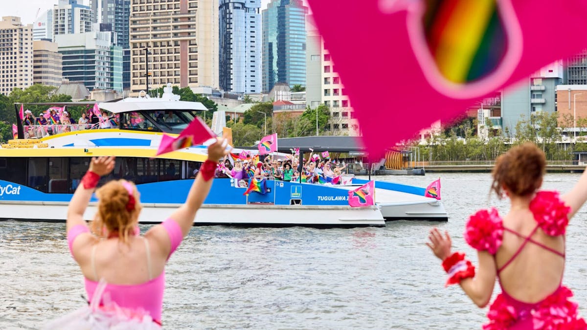 Two people in vibrant pink outfits cheer as a CityCat with colorful flags passes by on the river.