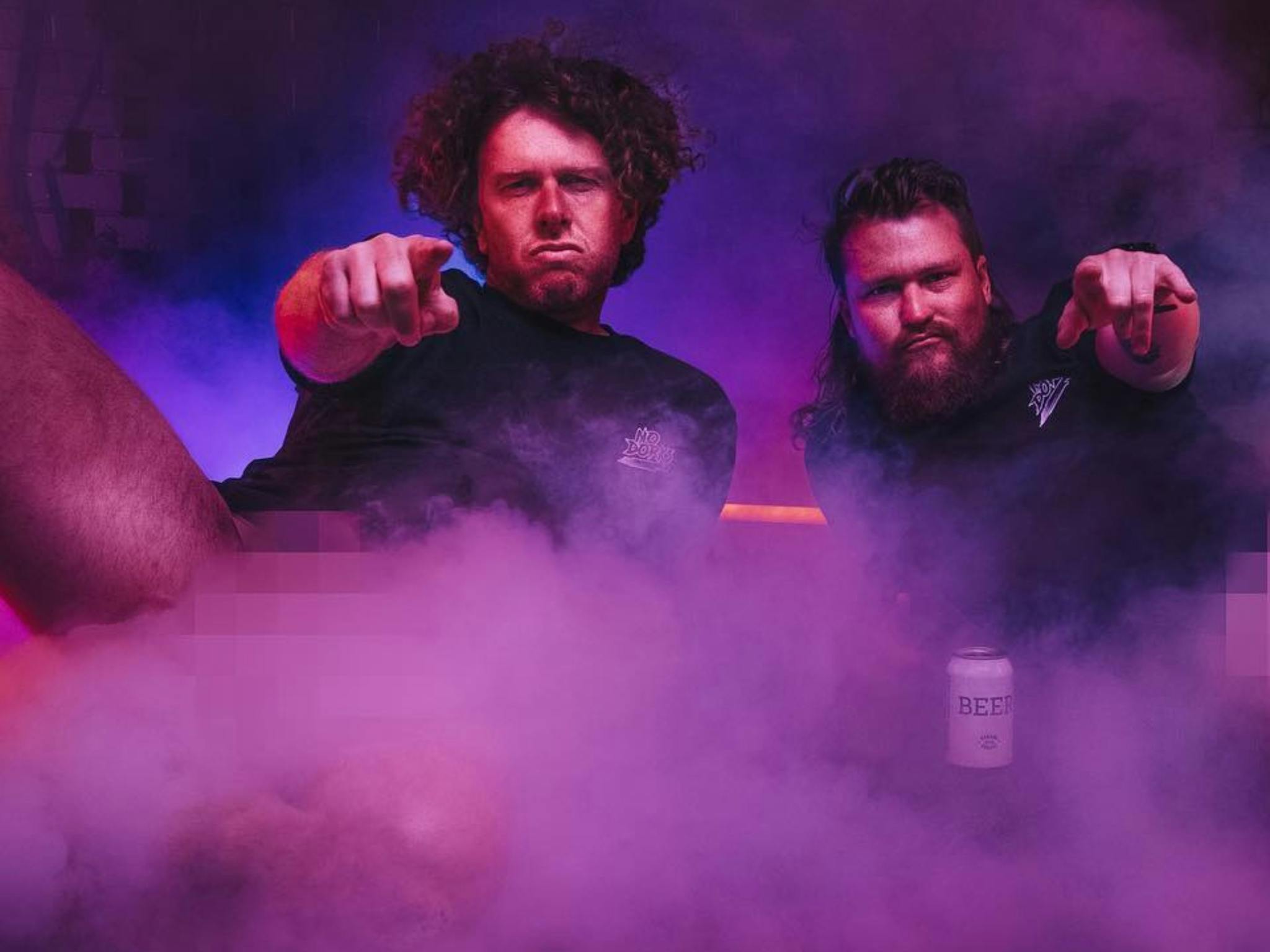 Brett and Nick sit in a purple smoky room. They are pointing at the camera.