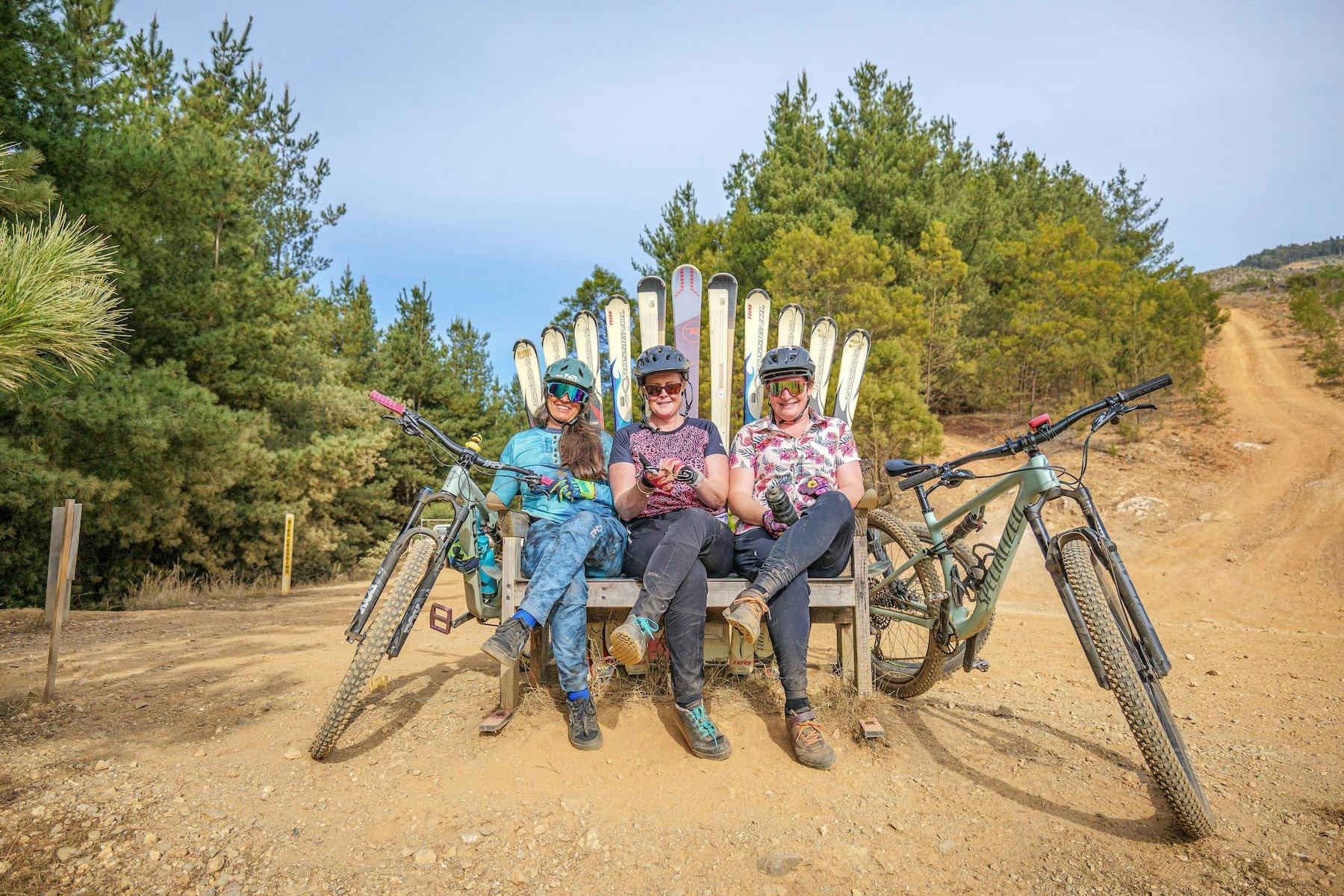 Three female mountain bikers sit on a seat made of skis with their bikes around them.