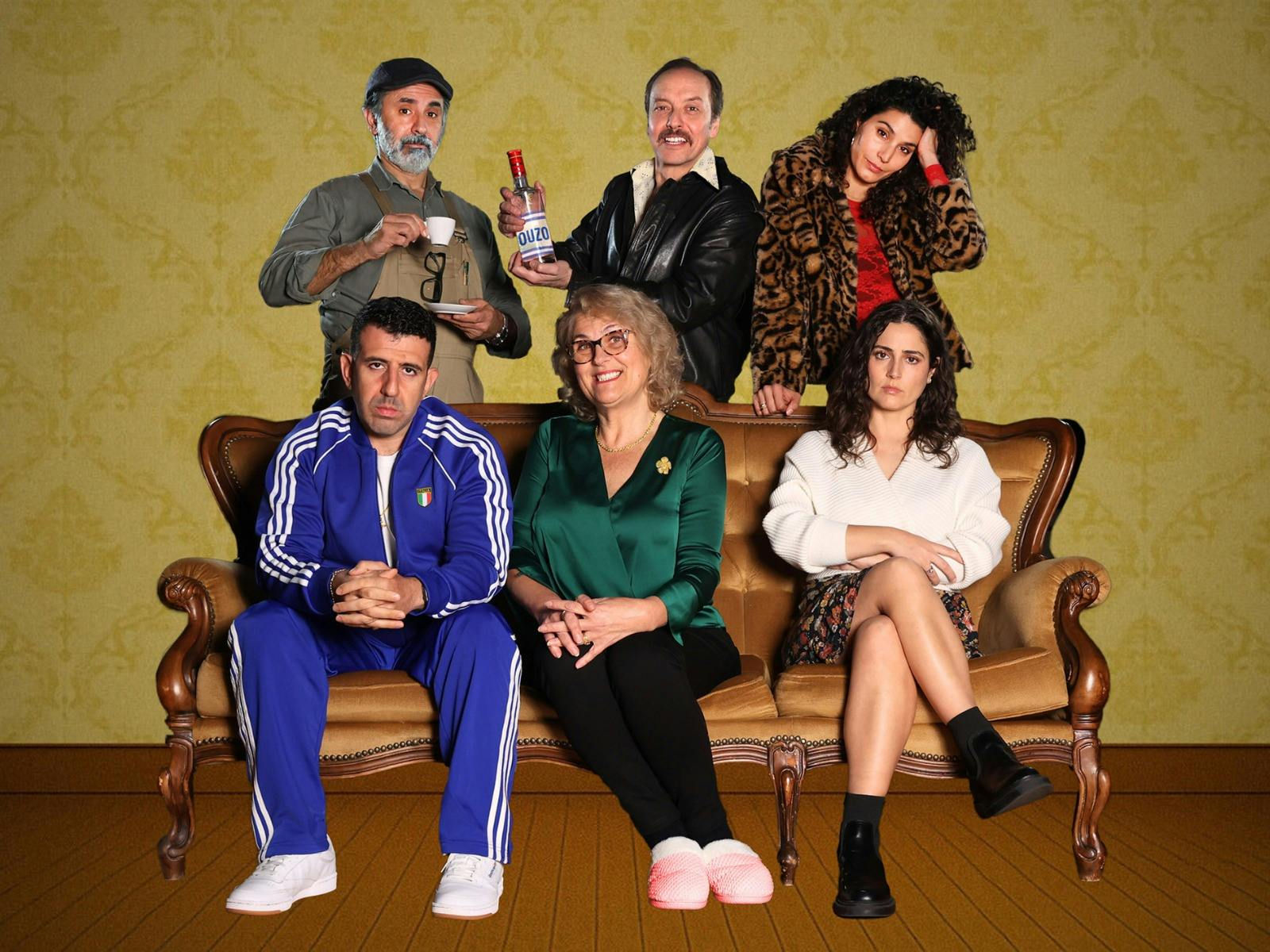 Six people pose in two rows on and behind a vintage sofa against a patterned yellow wall