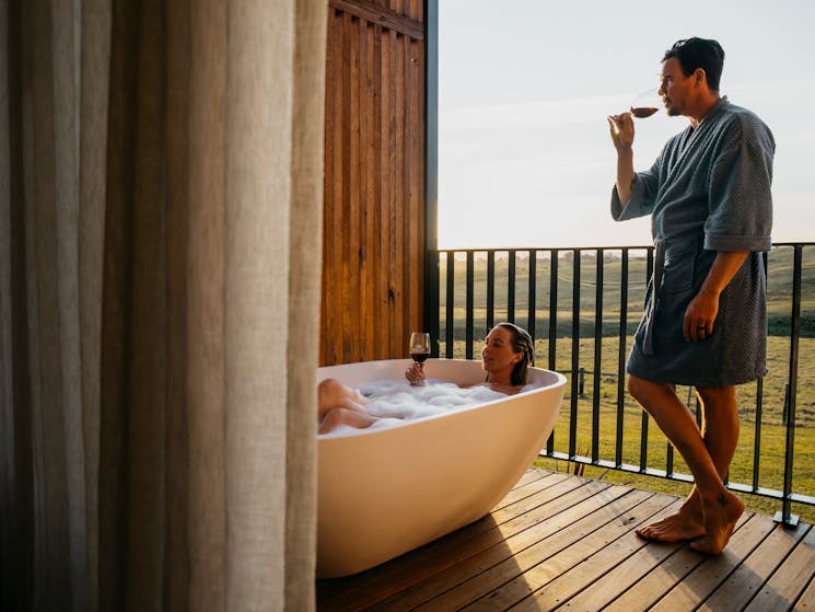 Woman enjoys outdoor bathtub drinking wine, man looking at the view drinking red wine.