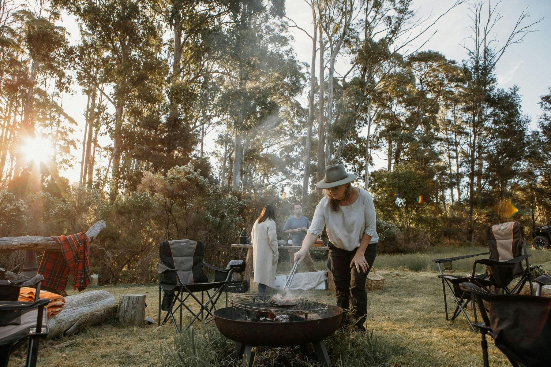 Bush camp setting woman wearing Akubra hat tending to fire with people standing around high table