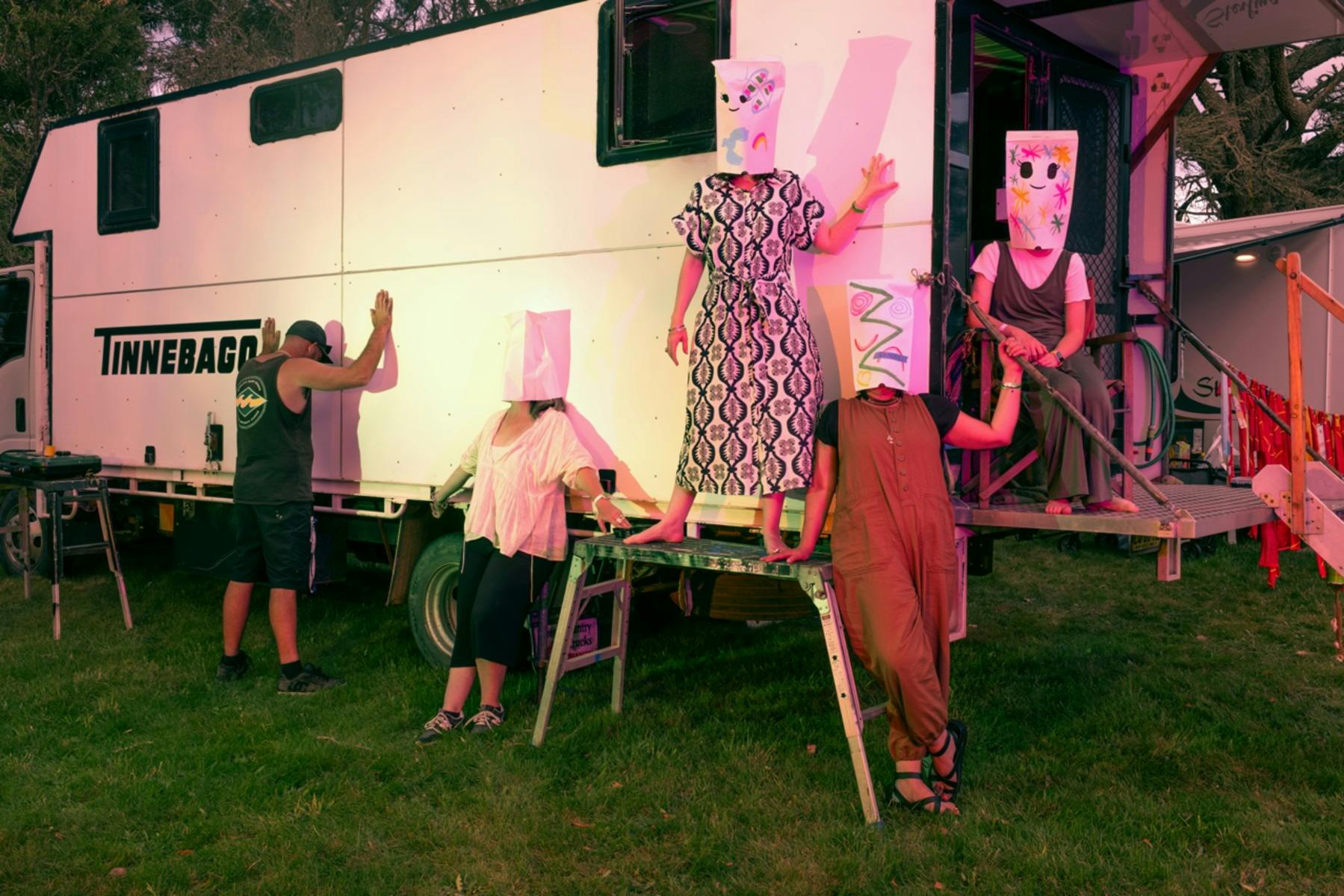 A photograph depicting five people wearing paper masks over their heads posing in front of a caravan