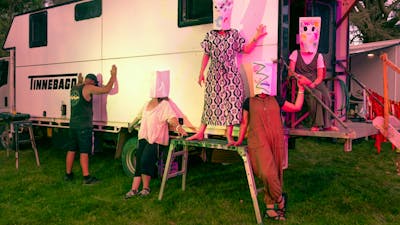 A photograph depicting five people wearing paper masks over their heads posing in front of a caravan