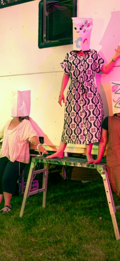 A photograph depicting five people wearing paper masks over their heads posing in front of a caravan