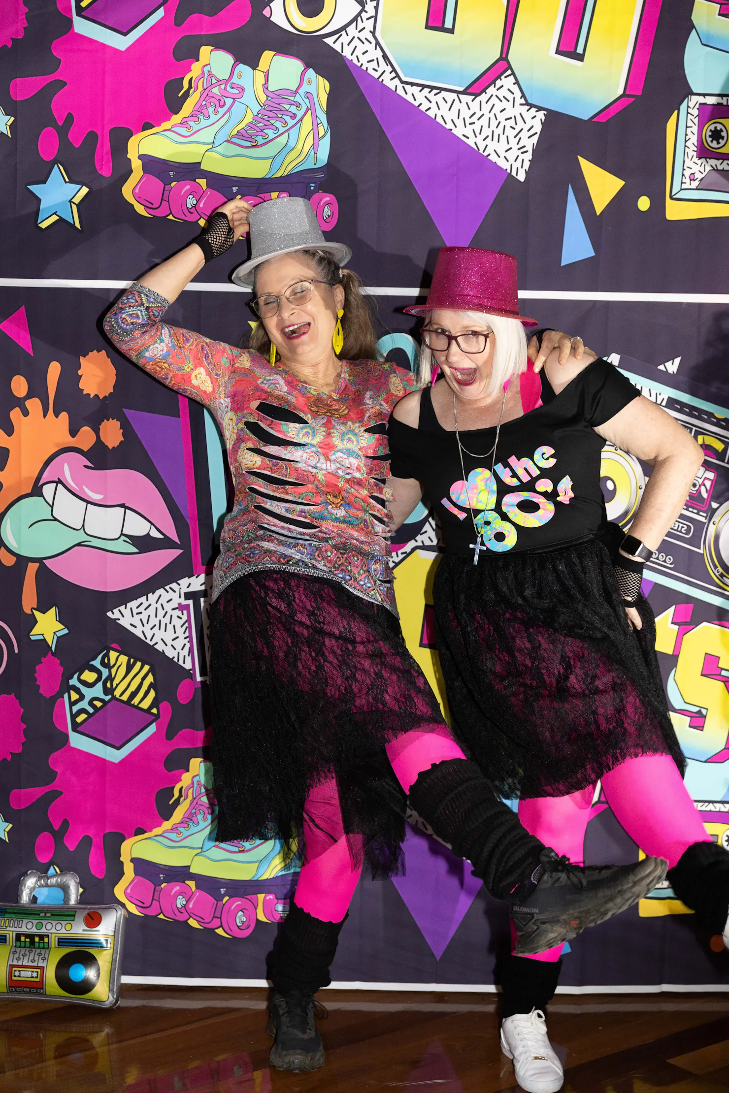 Two ladies pose and laugh in front of an 80s backdrop