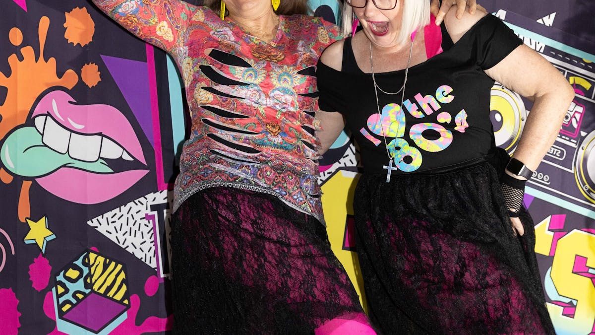 Two ladies pose and laugh in front of an 80s backdrop