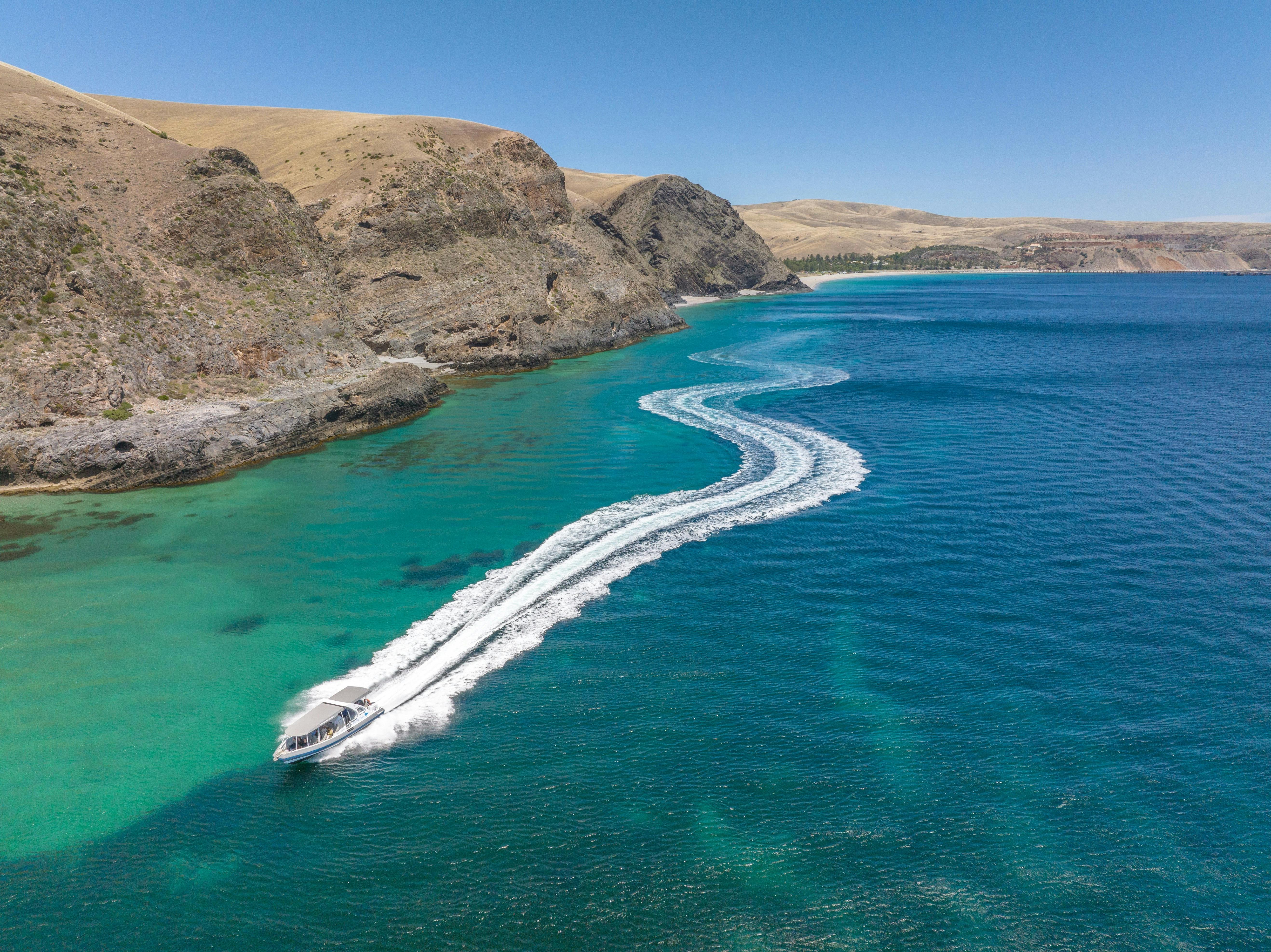 A boat speeds through clear turquoise water, leaving a wake near a rocky shoreline.