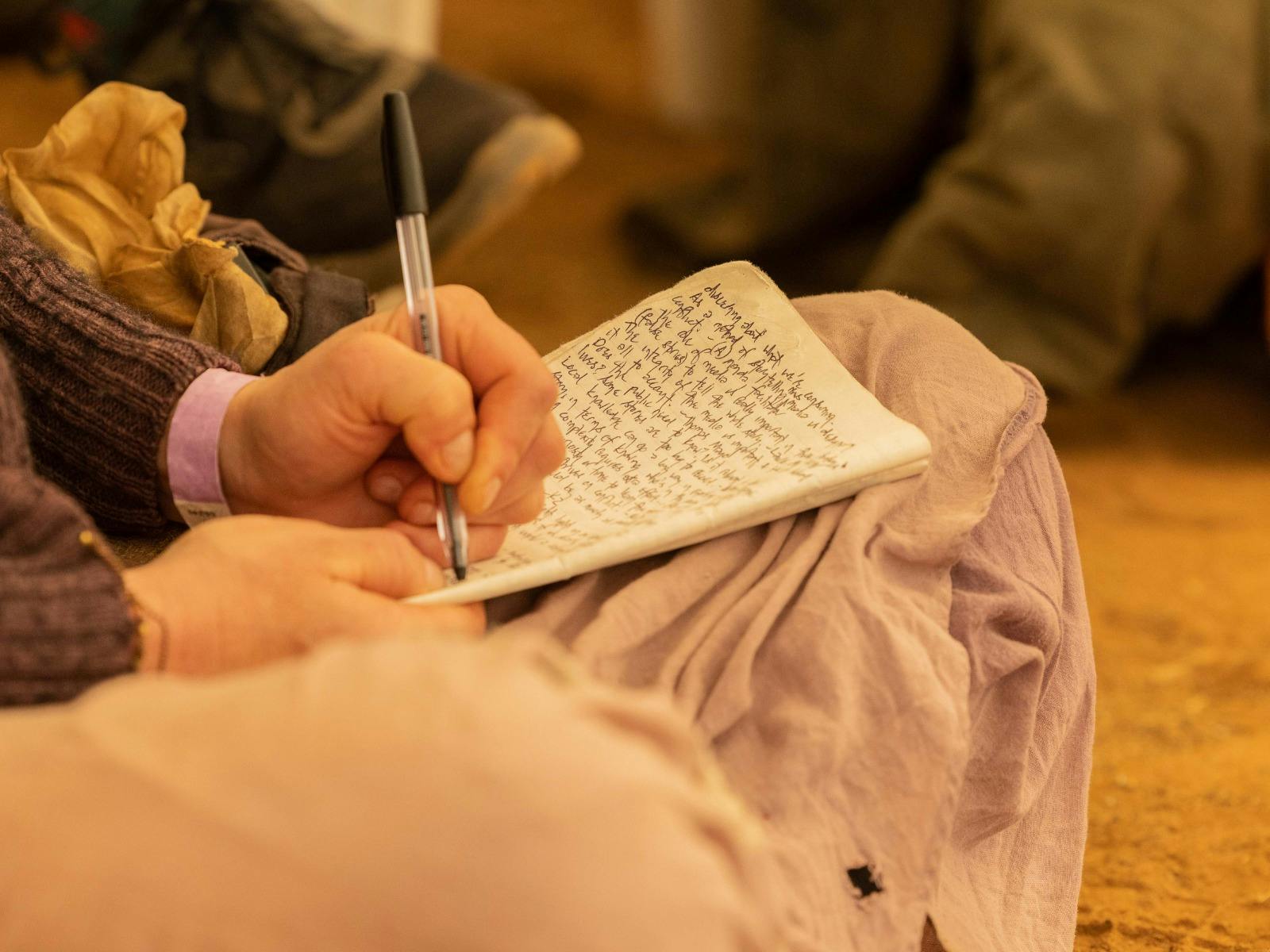 A person writes in a notebook with a black pen, sitting on the ground in an outdoor setting.