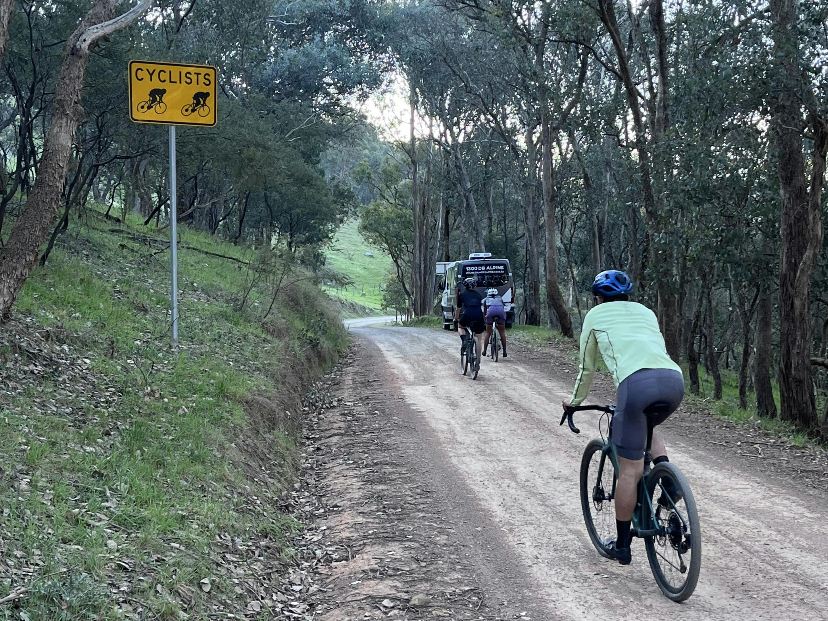 A group of cylists ride a gravel road toward Double Black Alpine vehicle ready for their shuttle.