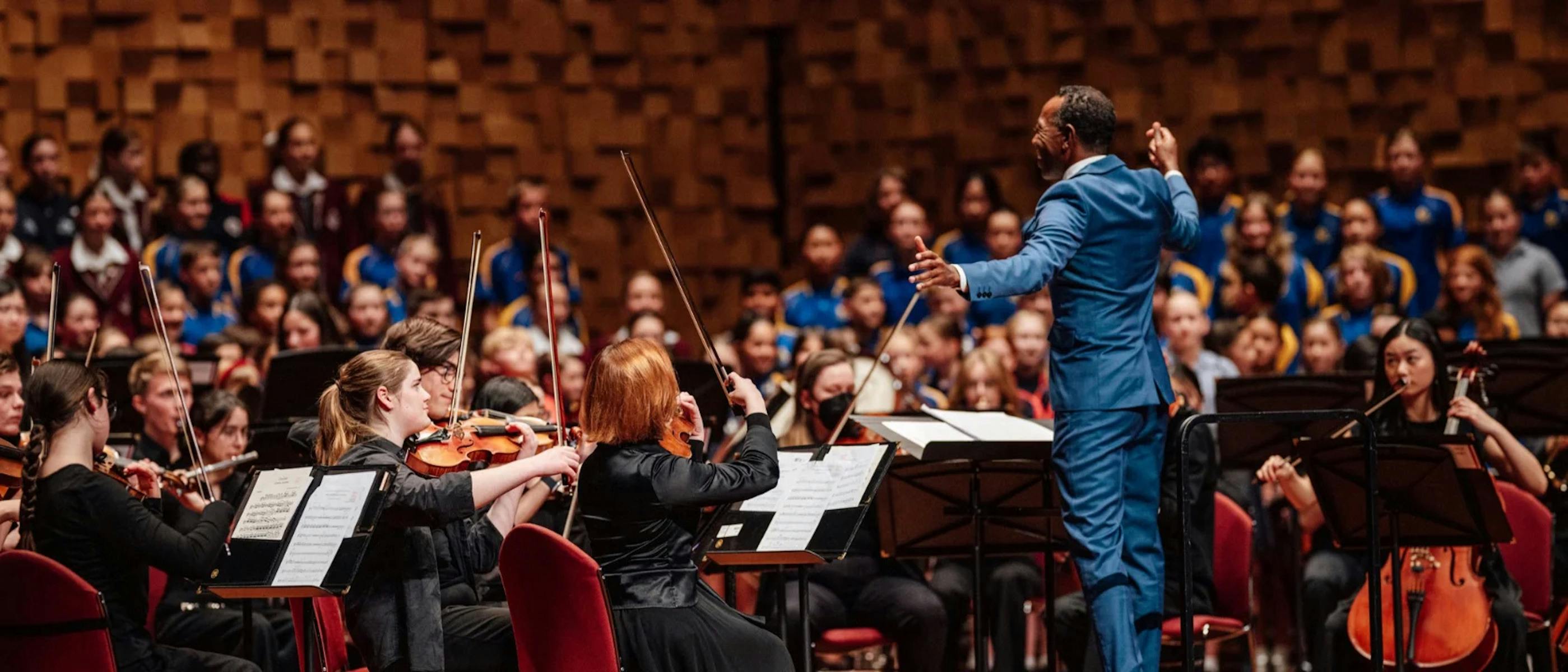 A conductor conducts an orchestra and a group of children singing