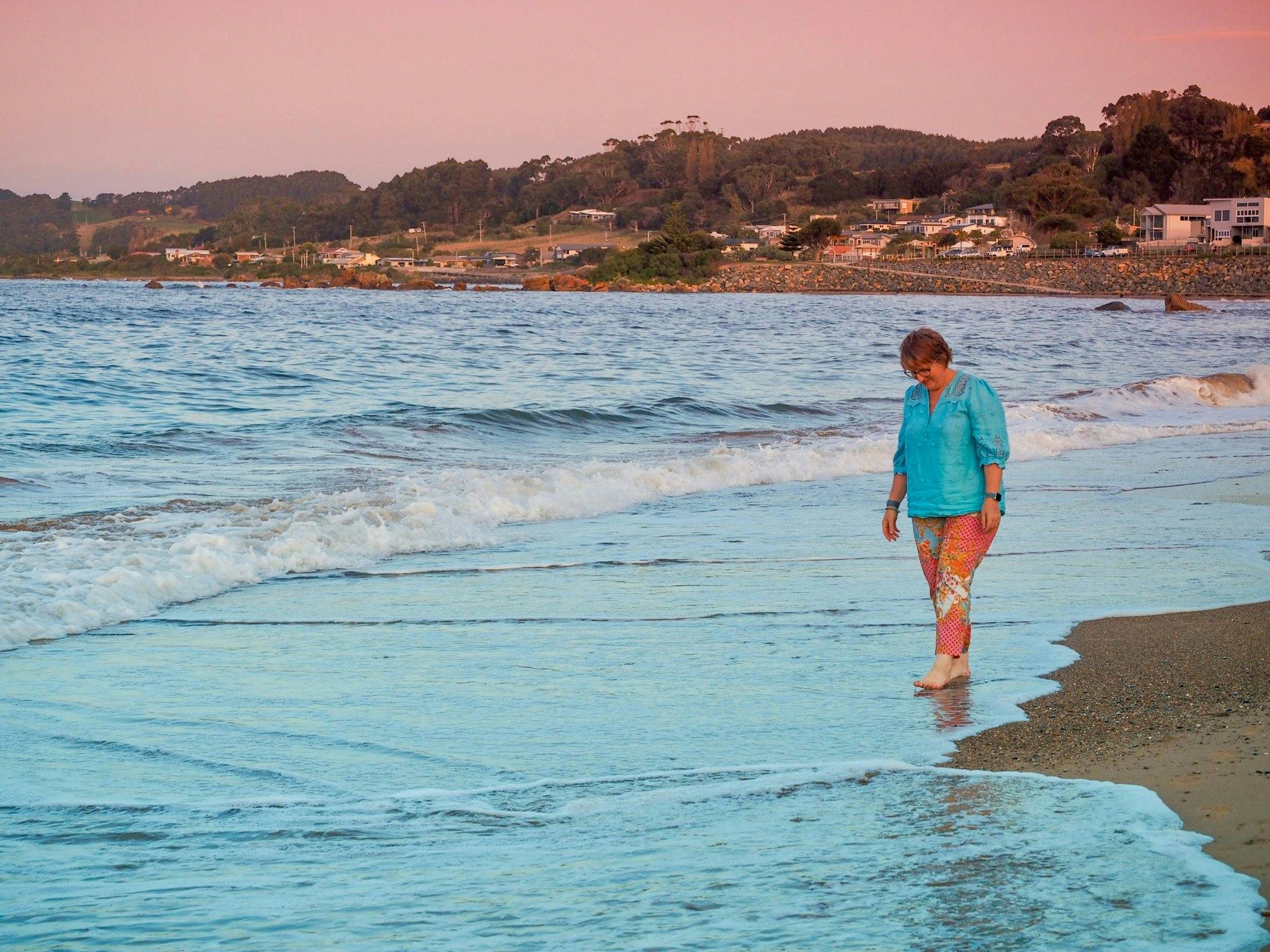 Woman walking barefoot on Penguin beach, dipping toes into the water on edge of small waves.