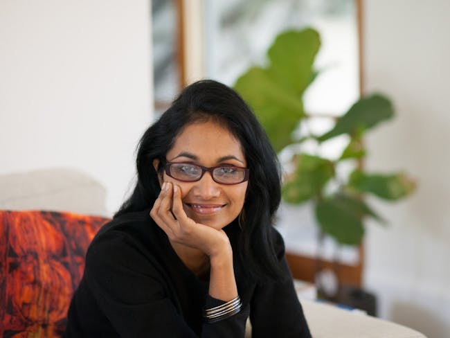 A dark-haired woman wearing glasses with a plant in the background