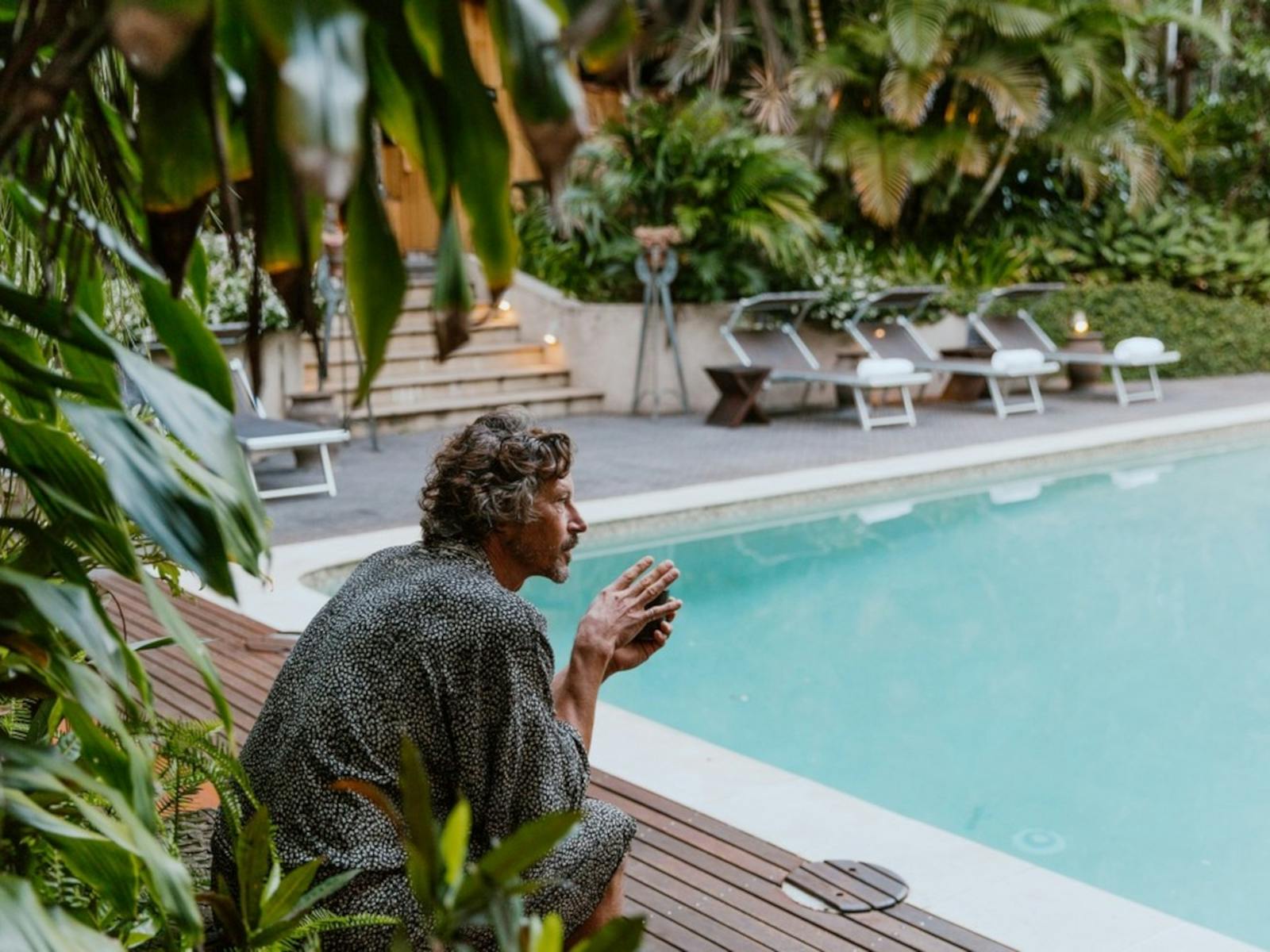 a man sits poolside wearing a robe and holding a warm drink