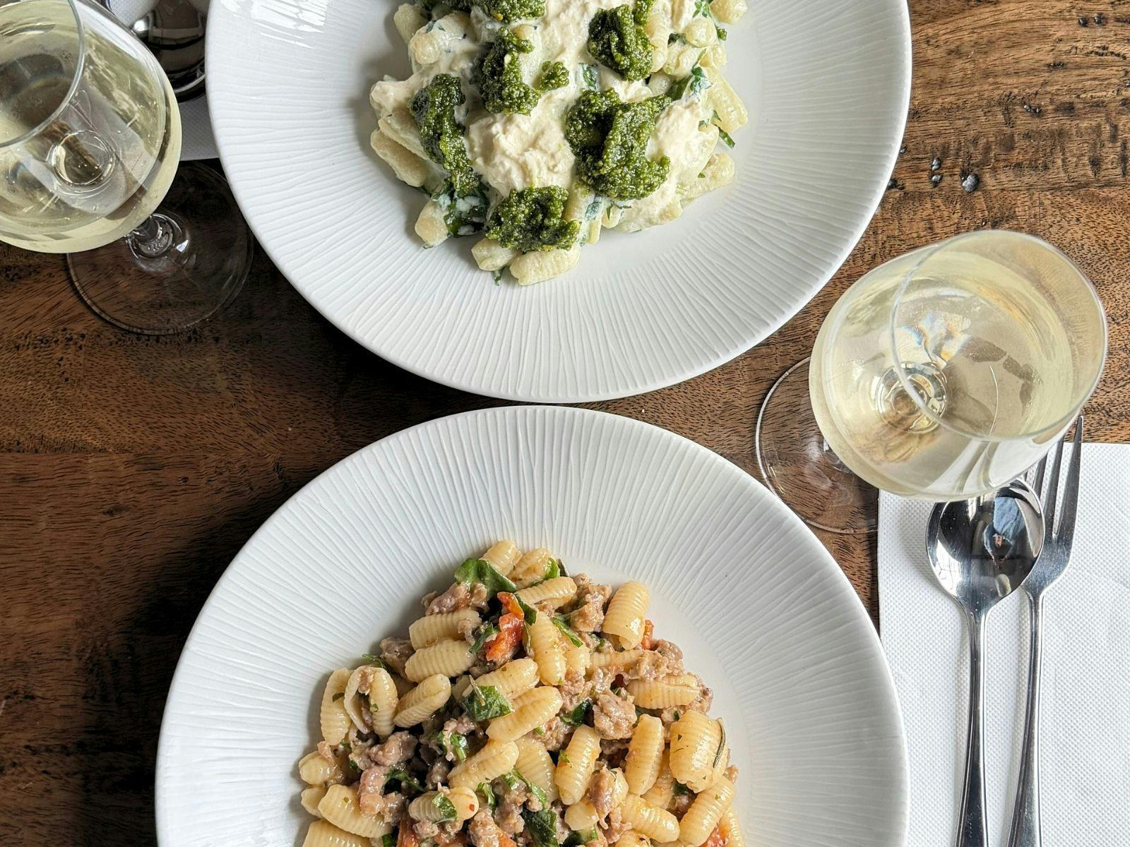 two plates of pasta on a dark timber table with two glasses of white wine