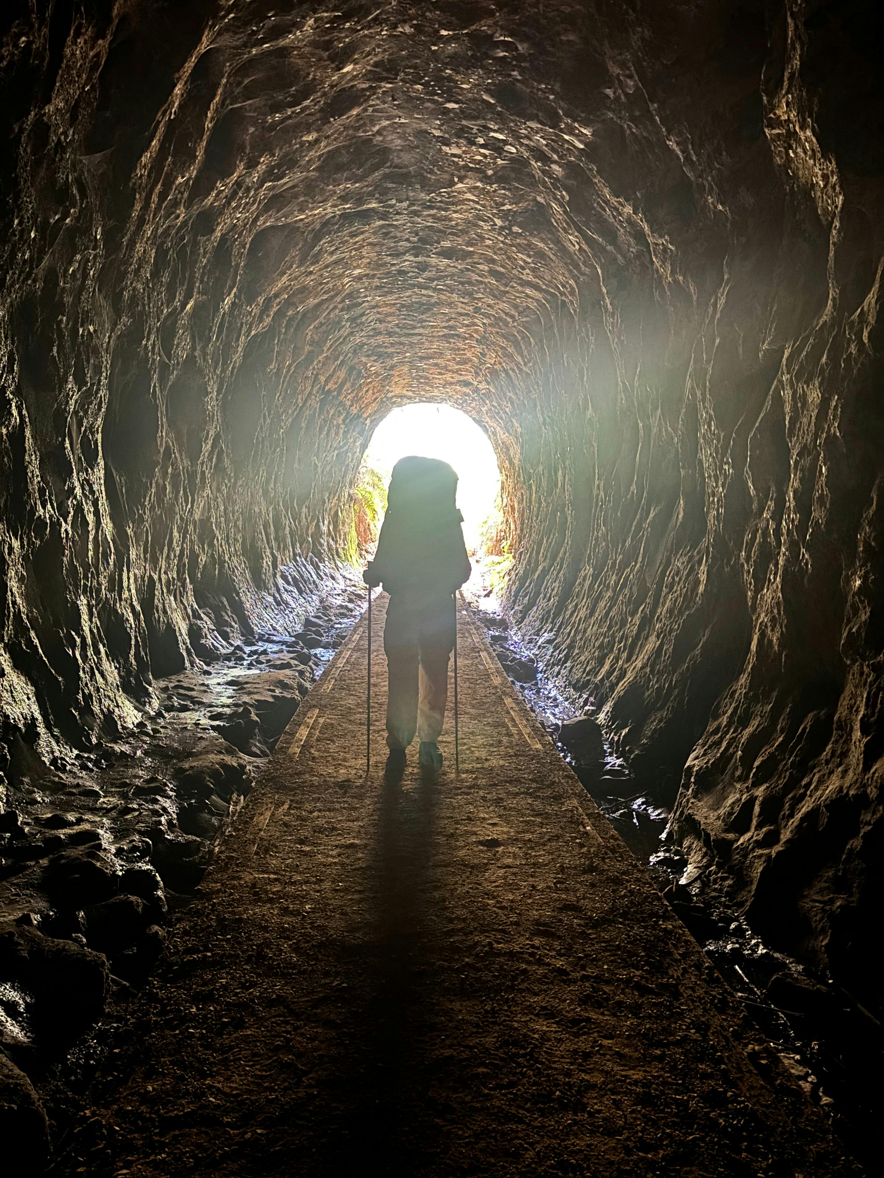 A dark figure walking along a disused railway tunnel, silhouetted by the sun behind.