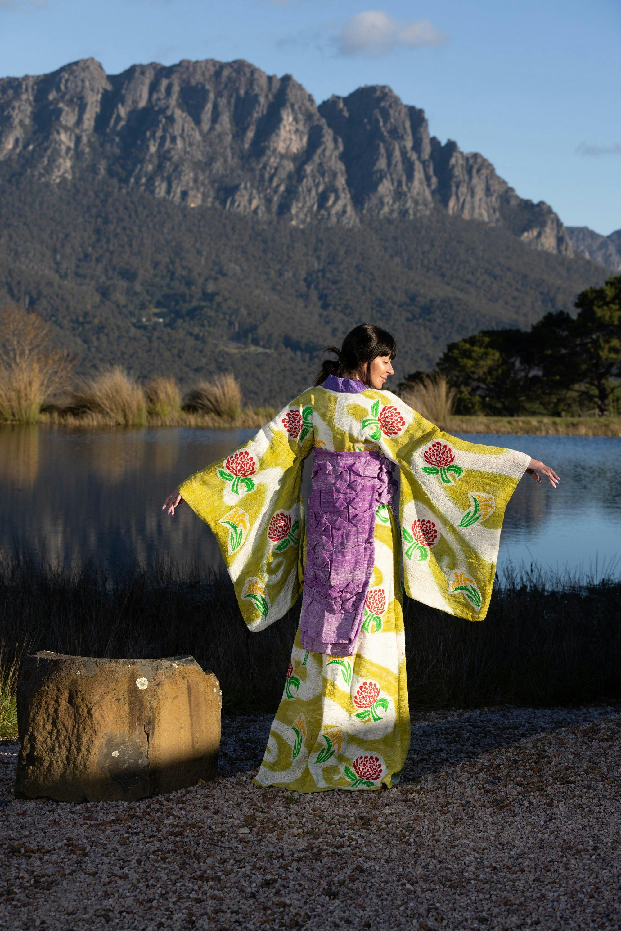 Model stands by a lake wearing a pale green kimono decorated with native flora.