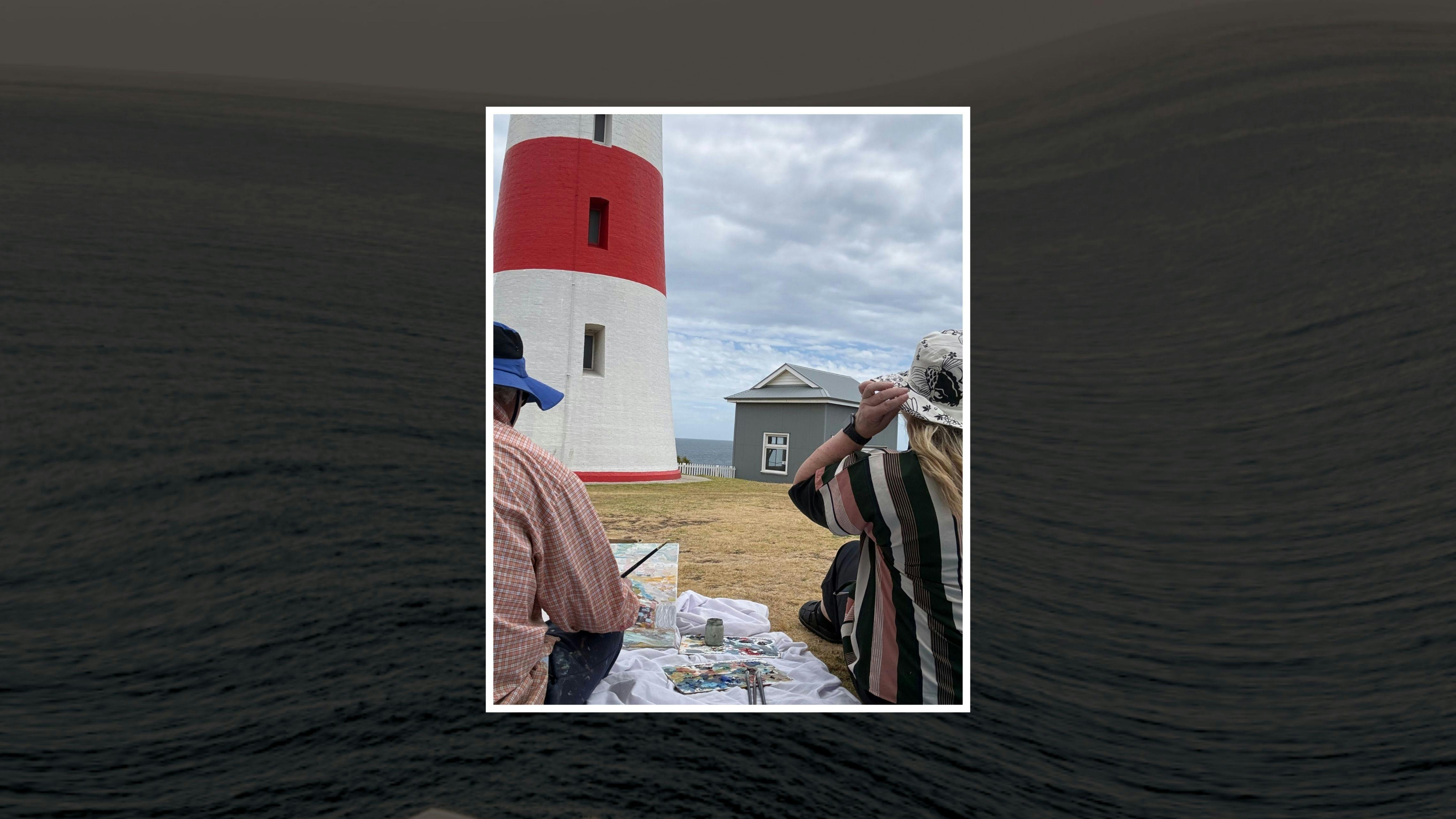Two artists creating near a red and white lighthouse by the sea under cloudy skies.