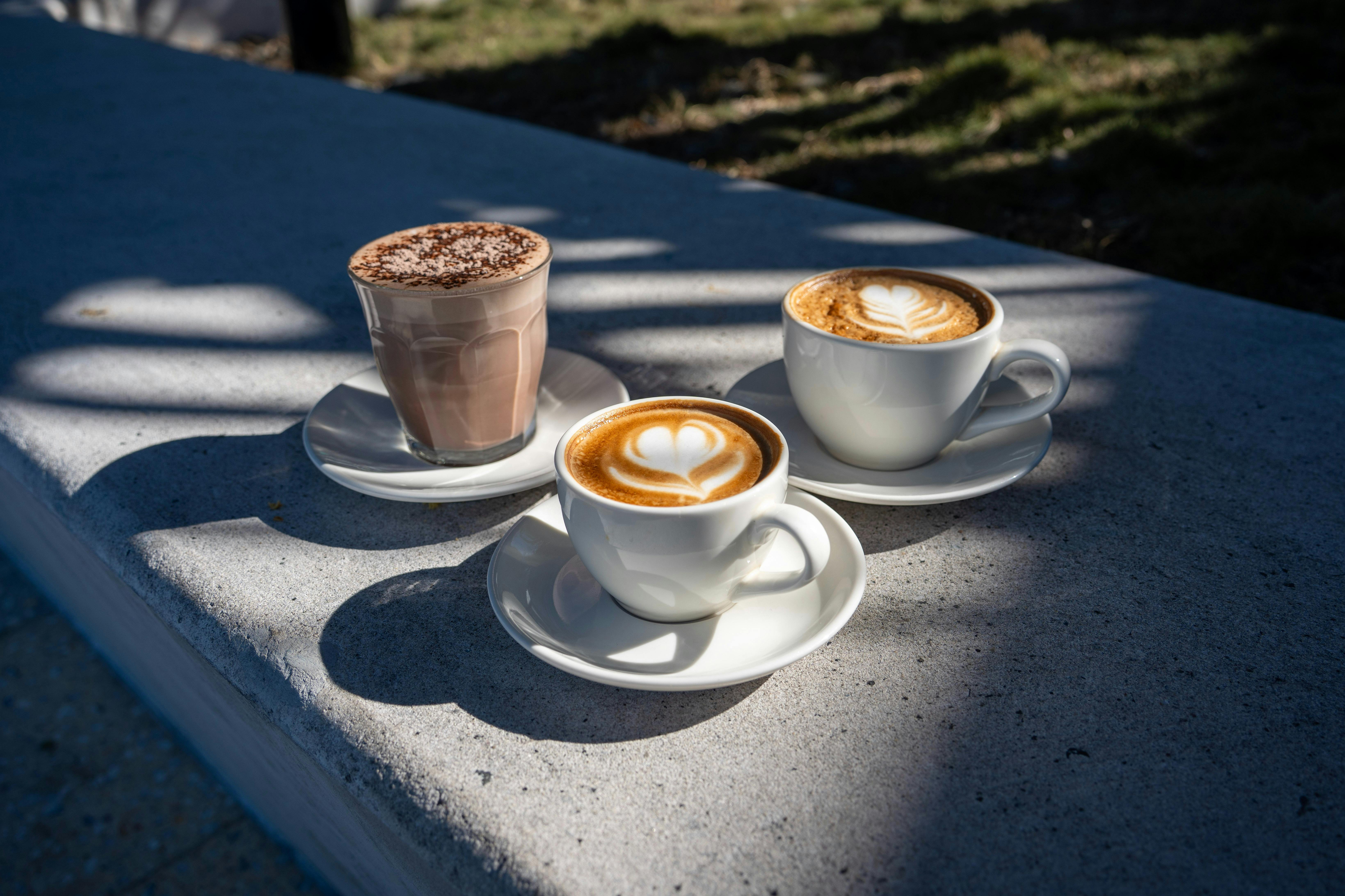 Three hot drinks, including coffees with latte art, on an outdoor café table.