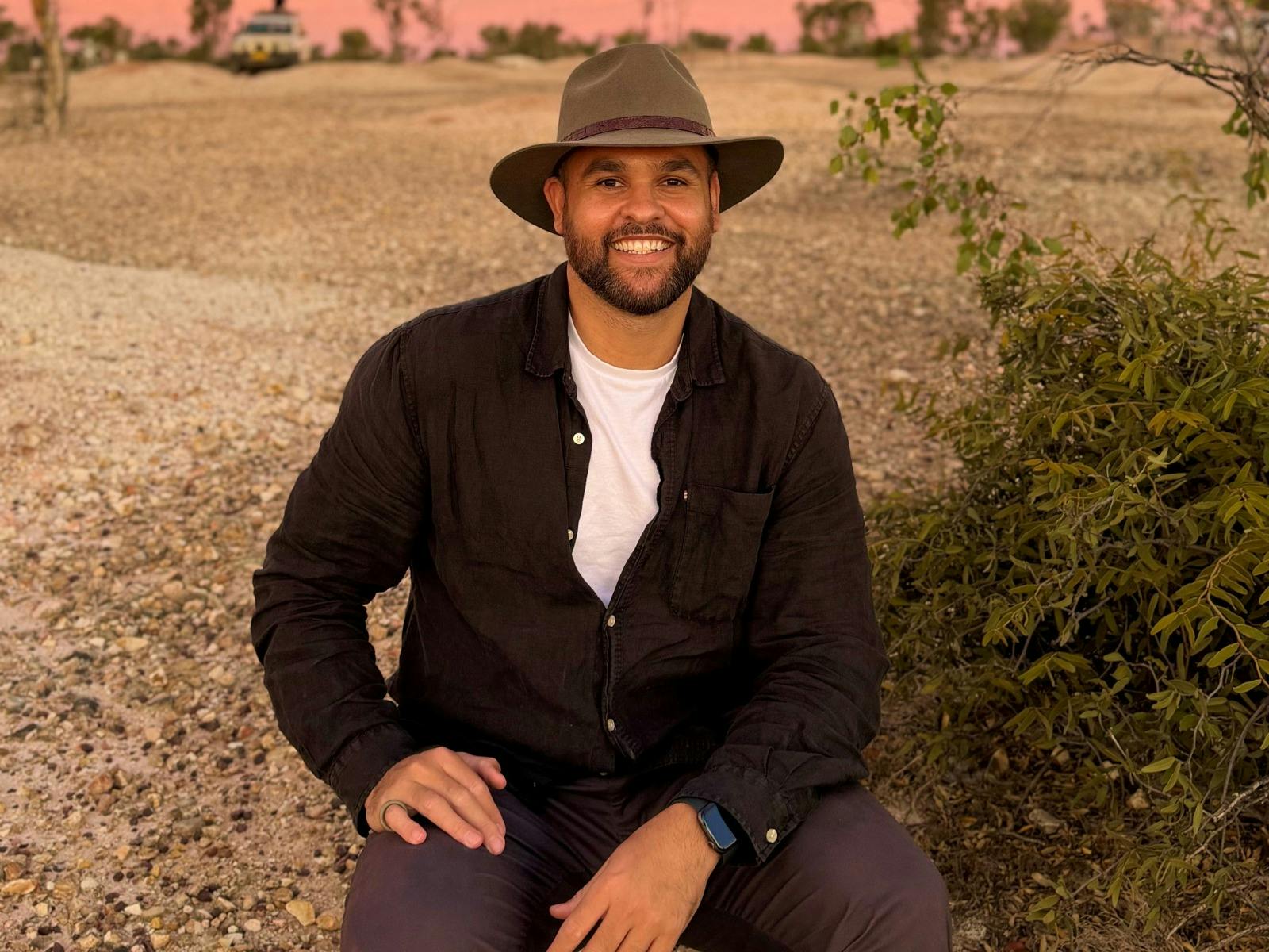 A smiling man with a beard and a wide-brimmed hat sits on a gravelly path in a dry, bushy landscape.