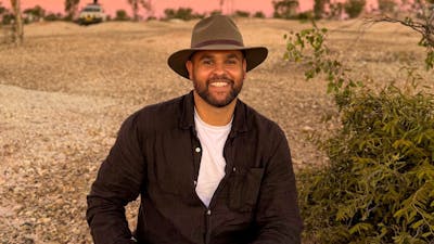 A smiling man with a beard and a wide-brimmed hat sits on a gravelly path in a dry, bushy landscape.