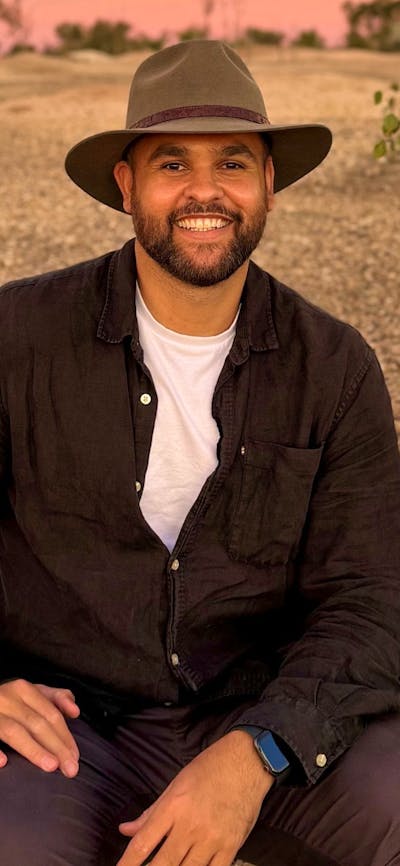 A smiling man with a beard and a wide-brimmed hat sits on a gravelly path in a dry, bushy landscape.
