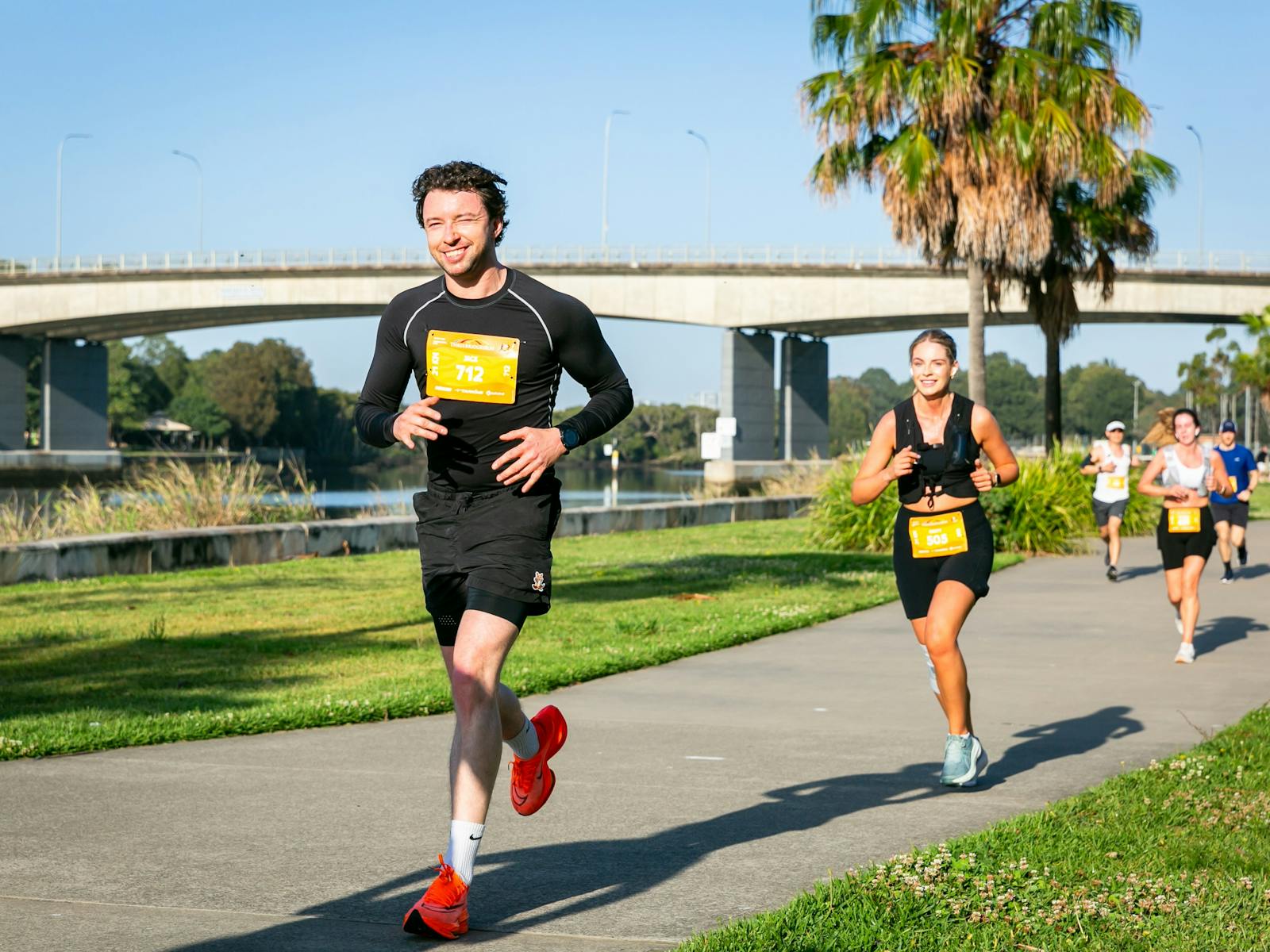 Two runners smiling running past a bridge along the Parramatta River
