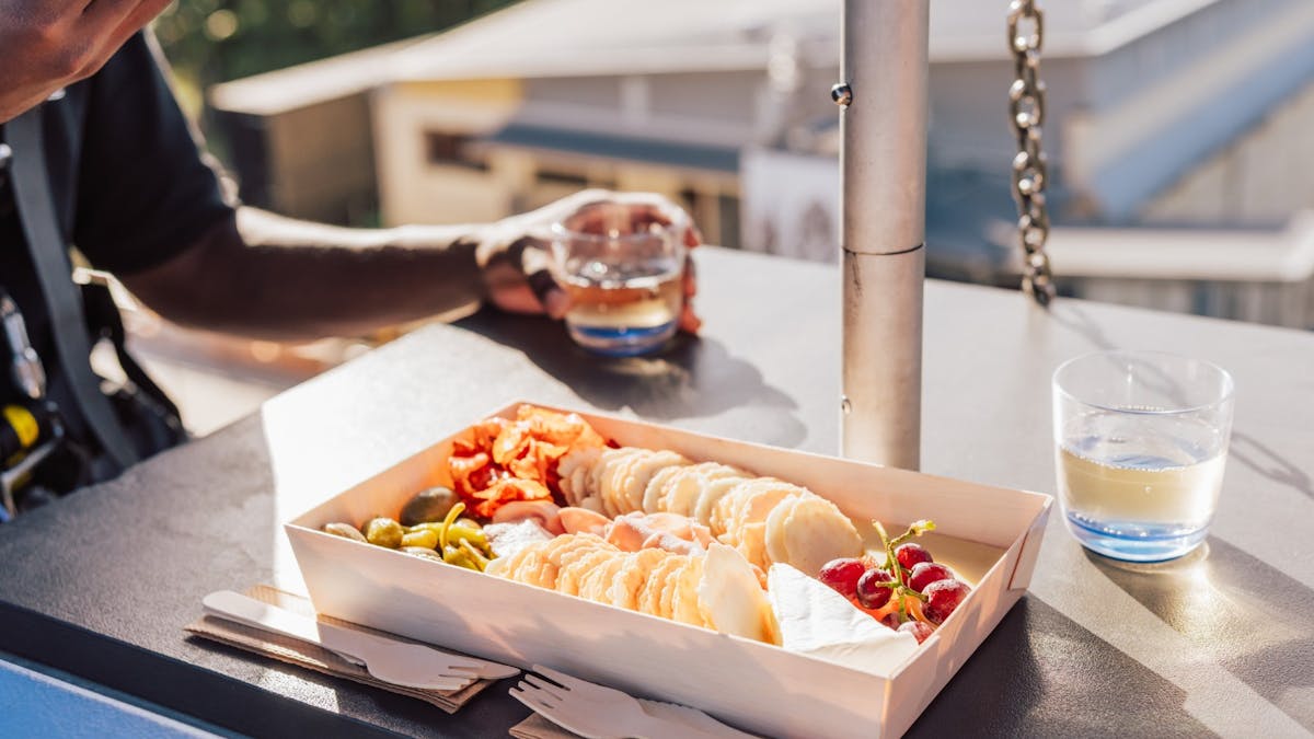 A box with assorted crackers, cheese, grapes, and olives on a table under sunlight.