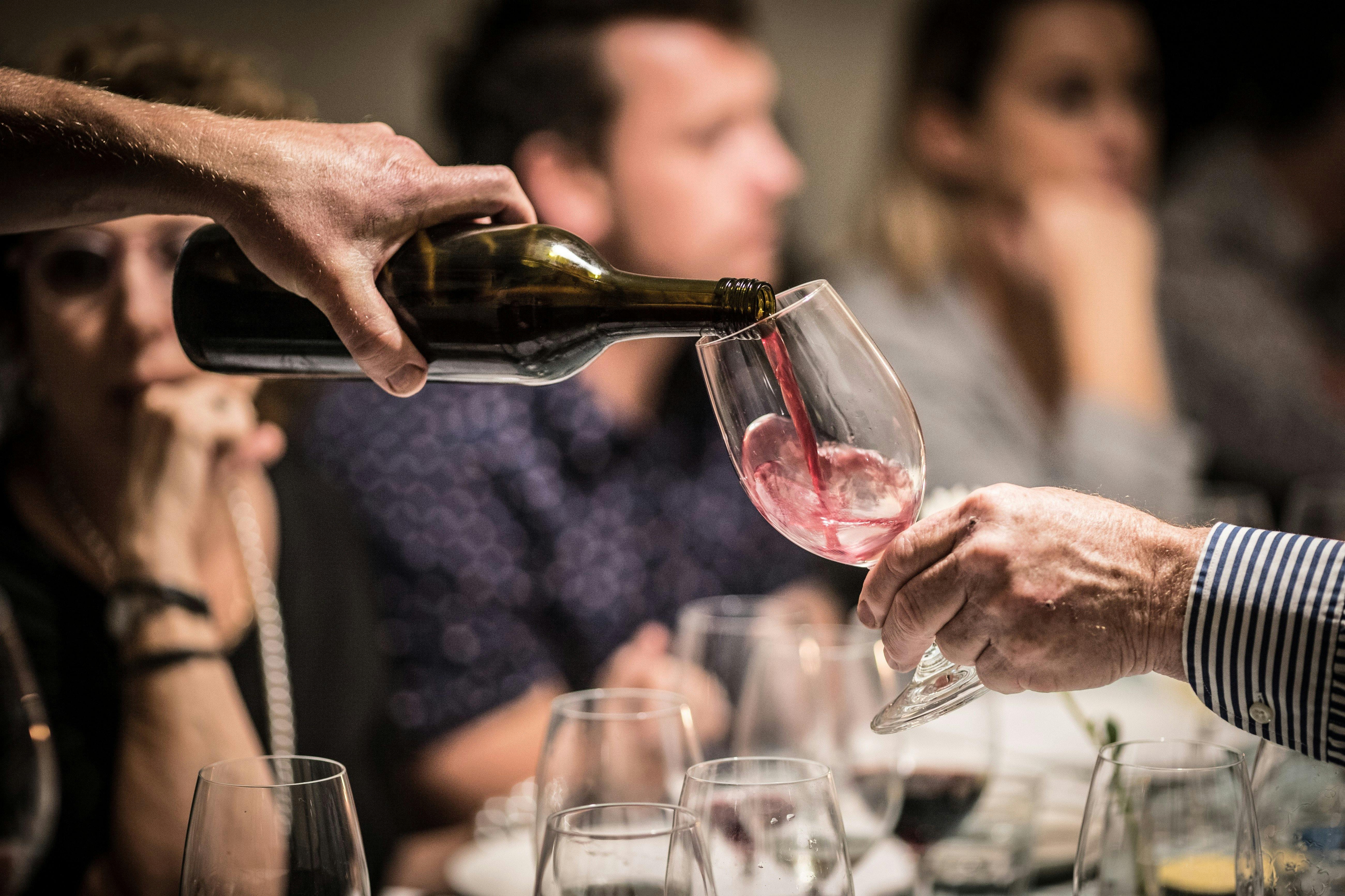 A glass of red wine is poured. In the background people are eating and smiling.