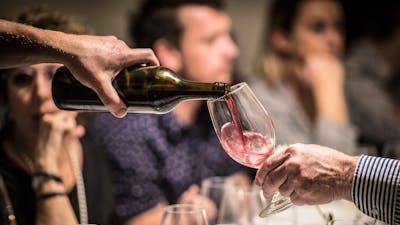 A glass of red wine is poured. In the background people are eating and smiling.