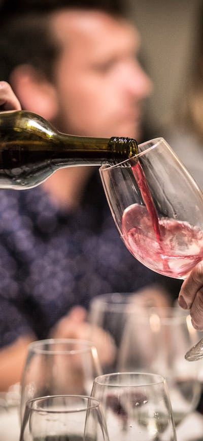 A glass of red wine is poured. In the background people are eating and smiling.