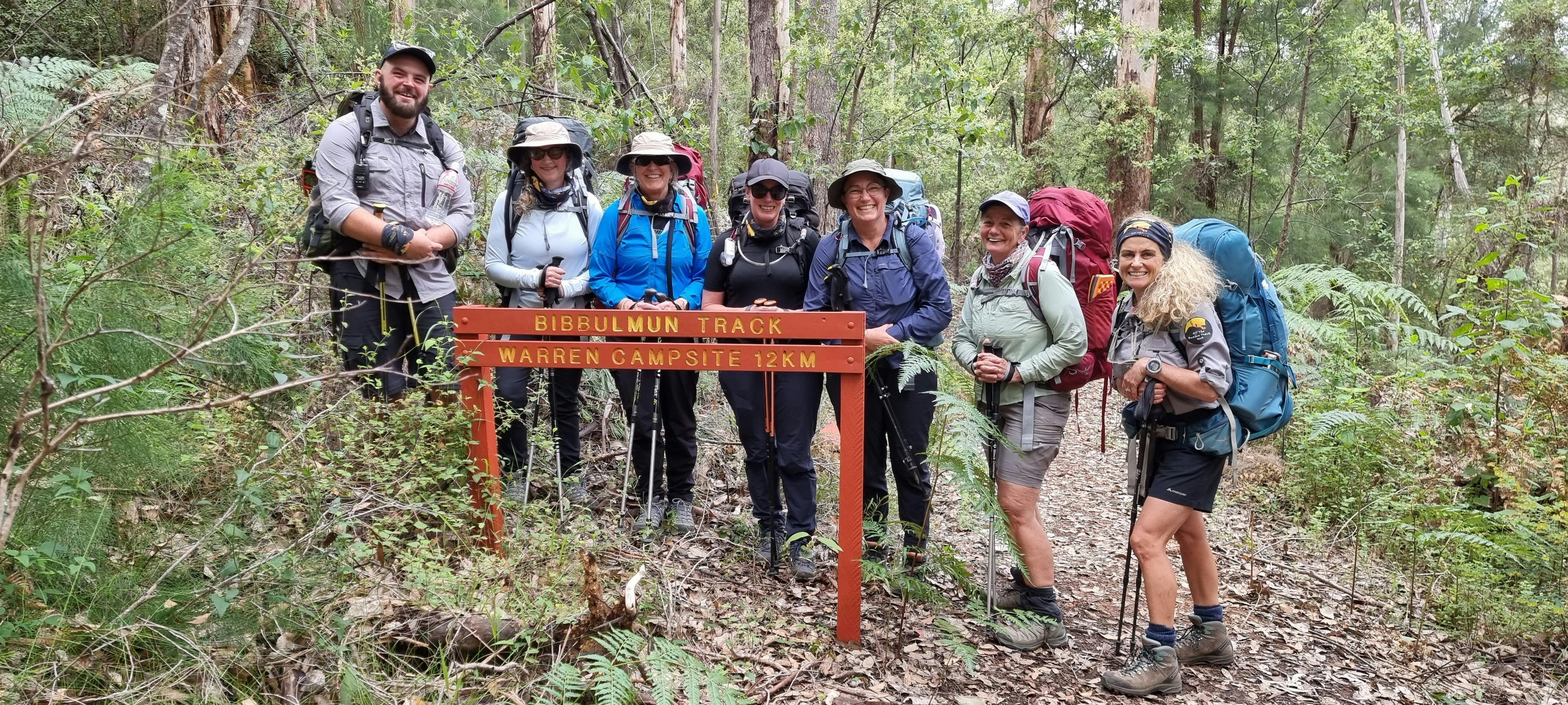 Hiking group of seven people smiling behind Bibbulmun Track trail sign
