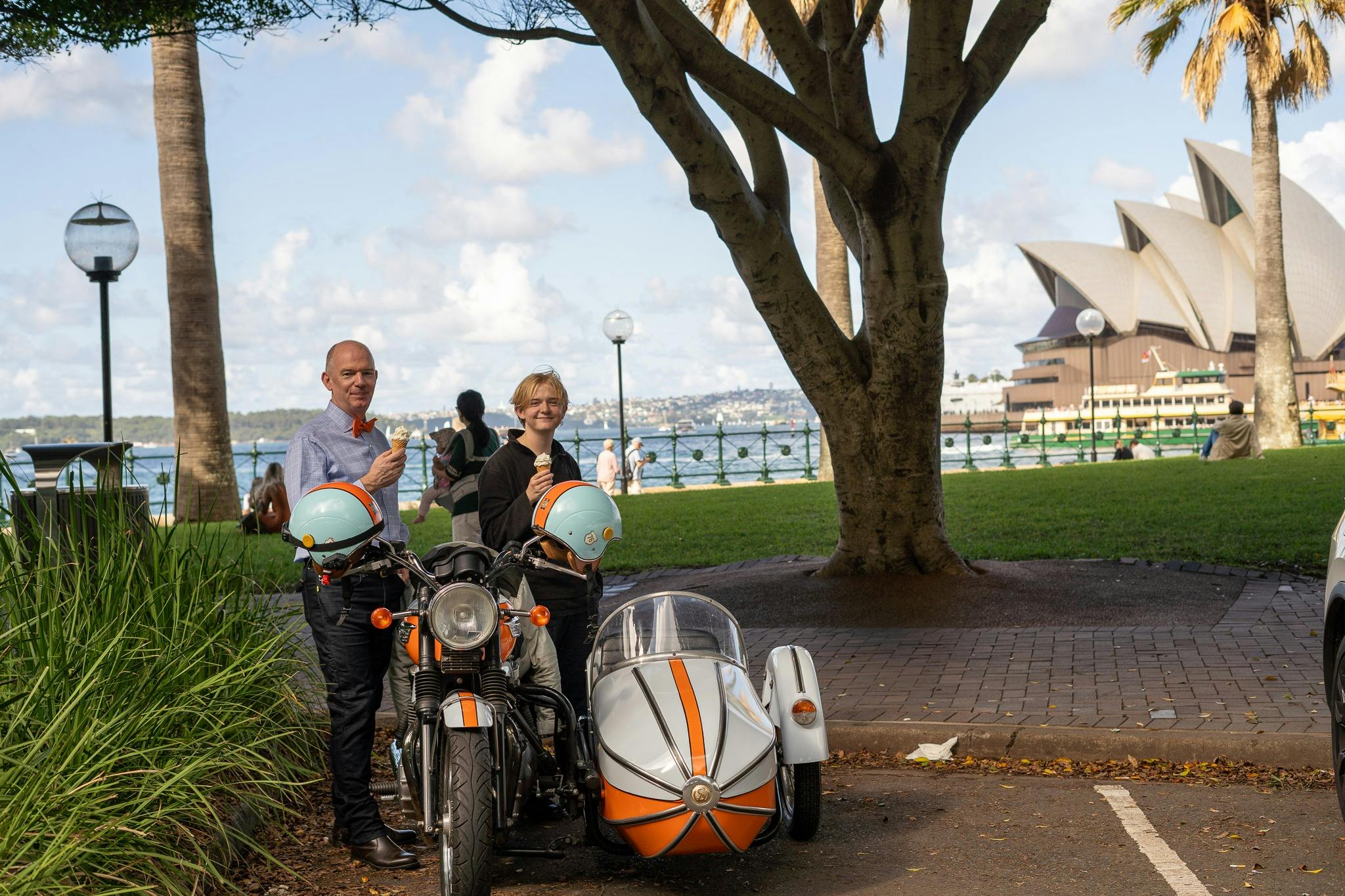 Two people eating icream by a motorcuyle and sidecar across the water from the Sydney Opera House