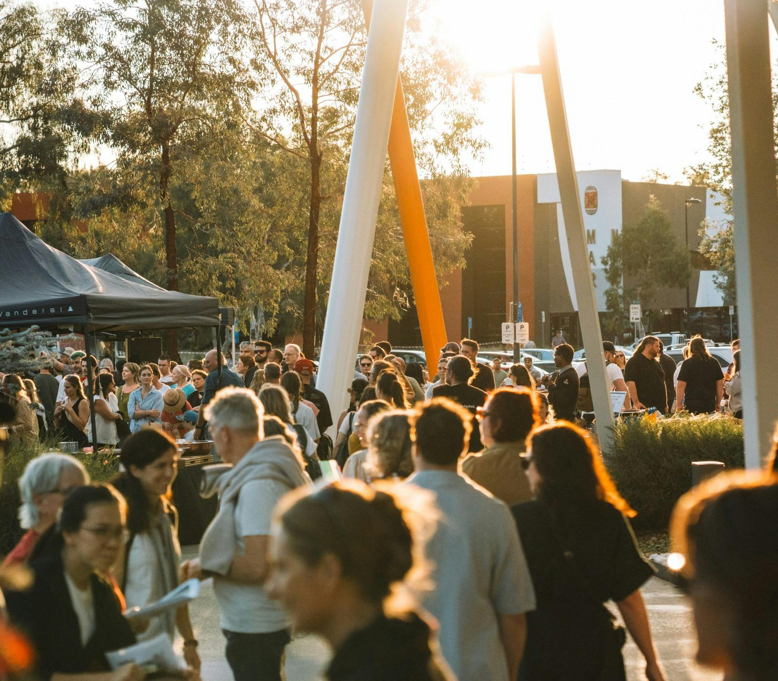 Bustling outdoor crowd at walking at sunset with marquees in the background.