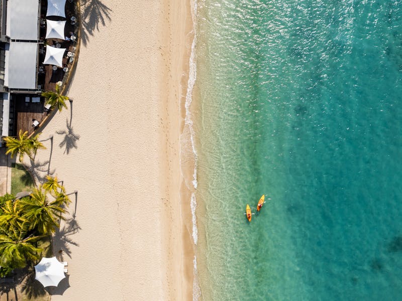 Aerial view of a sandy beach, turquoise water, palm trees, and two kayakers near the shoreline.