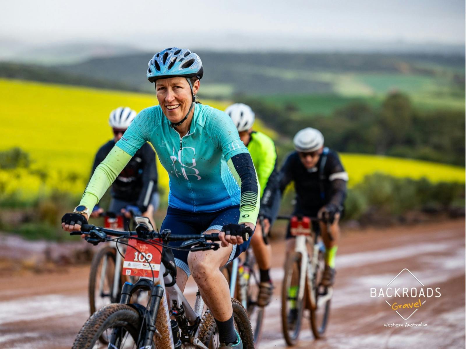 Cyclist riding a red dirt road with yellow canola fields in the background.