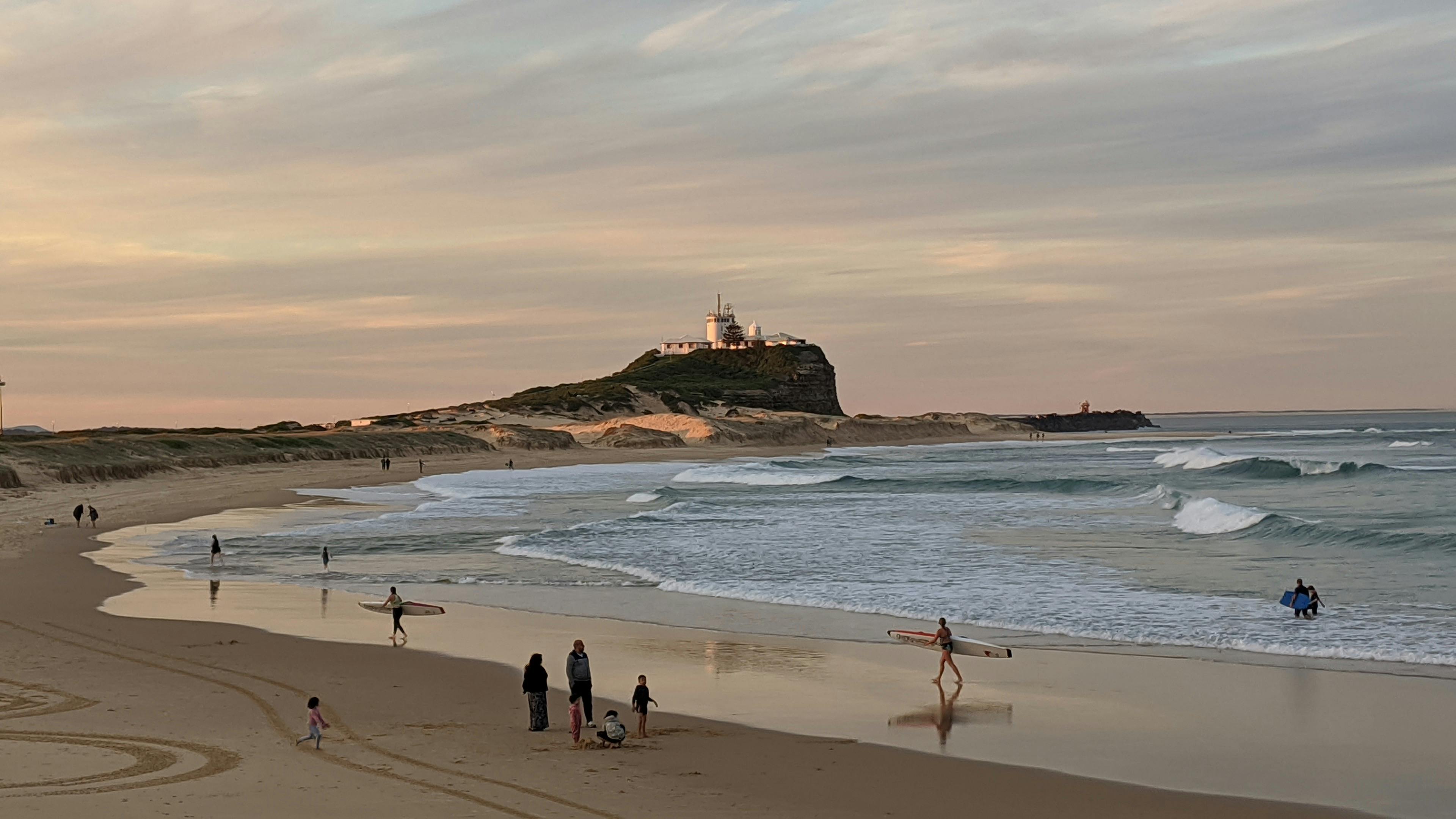 Photo of Nobbys Beach from the shore. People are in the sand. It is early evening.