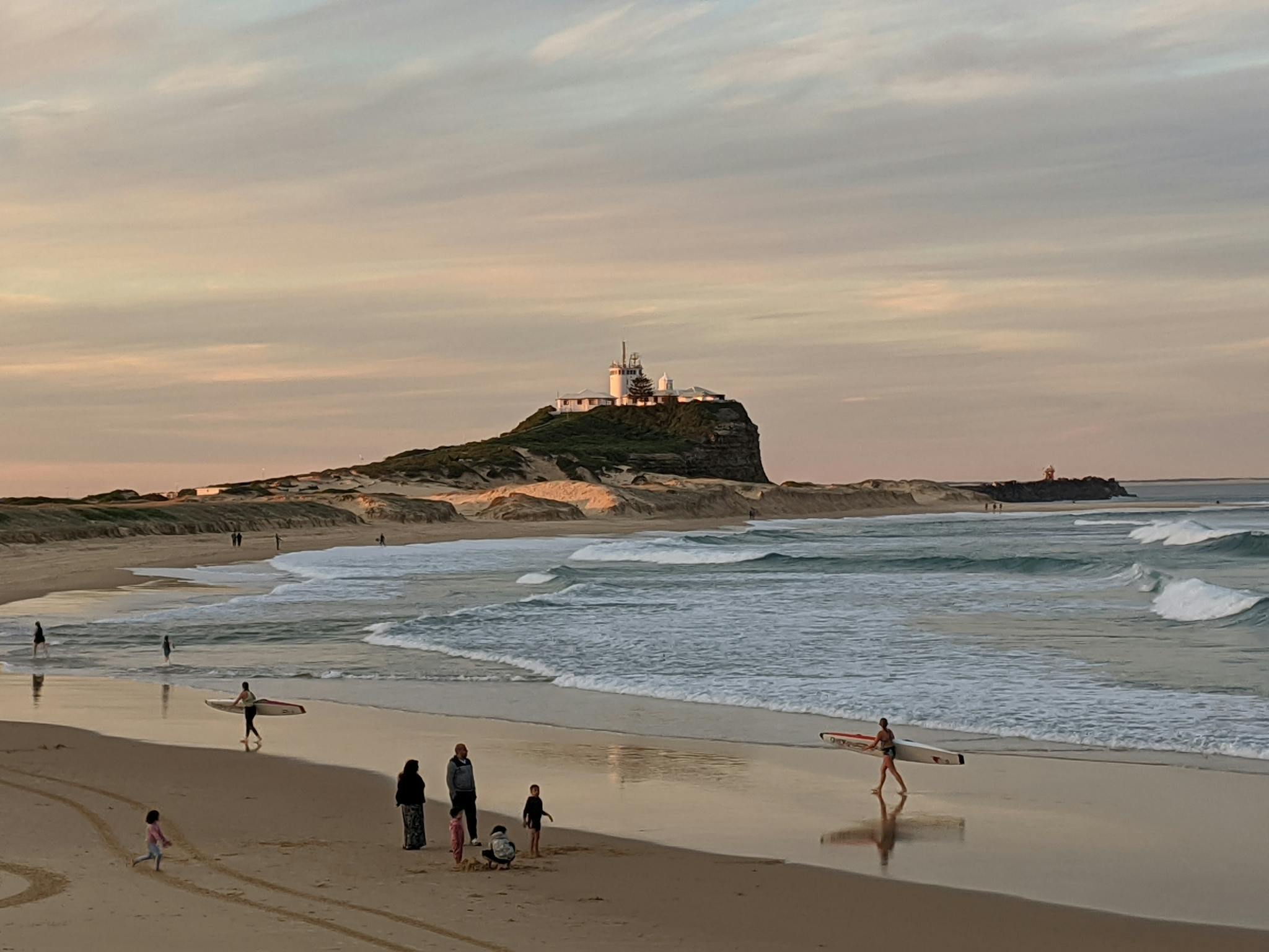 Photo of Nobbys Beach from the shore. People are in the sand. It is early evening.