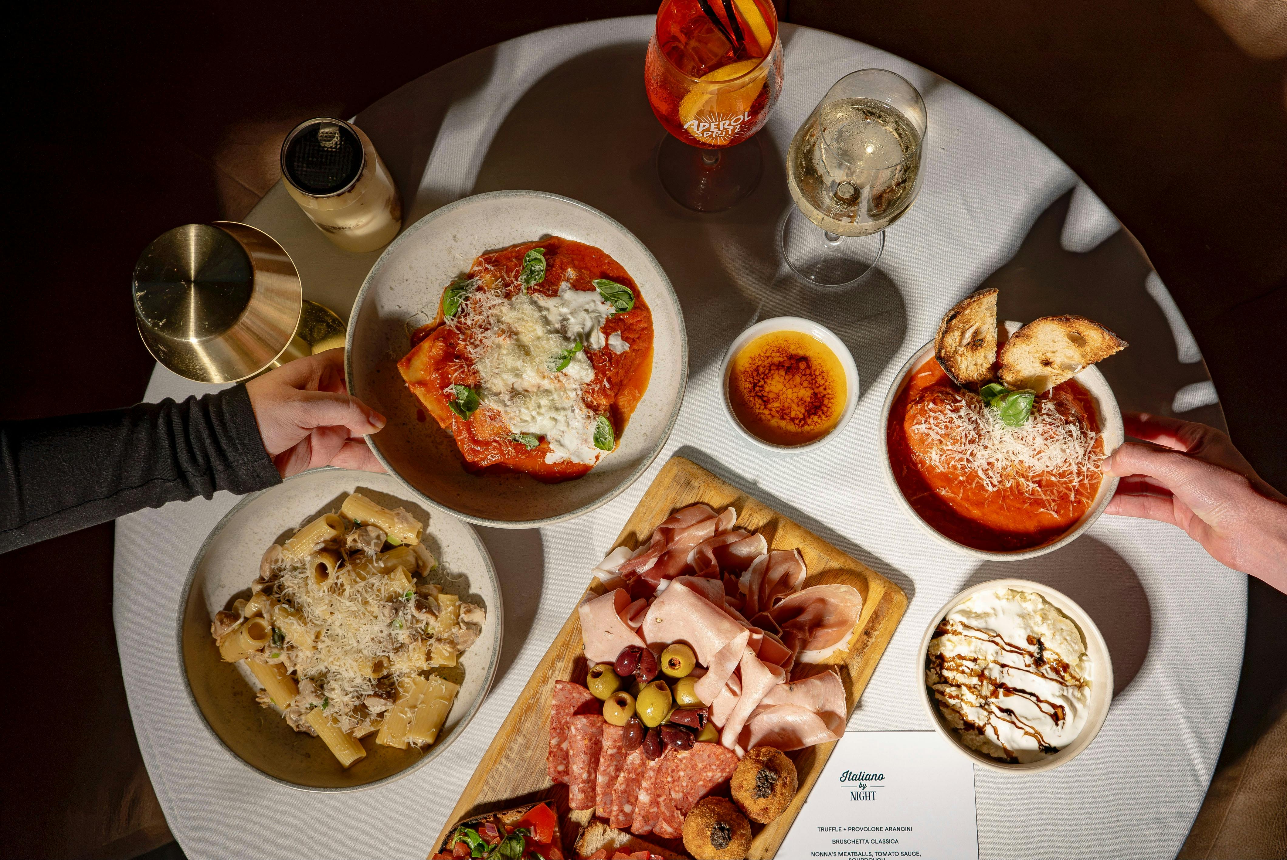 Friends share an Italian meal with pasta, soup, charcuterie, and drinks around a table.