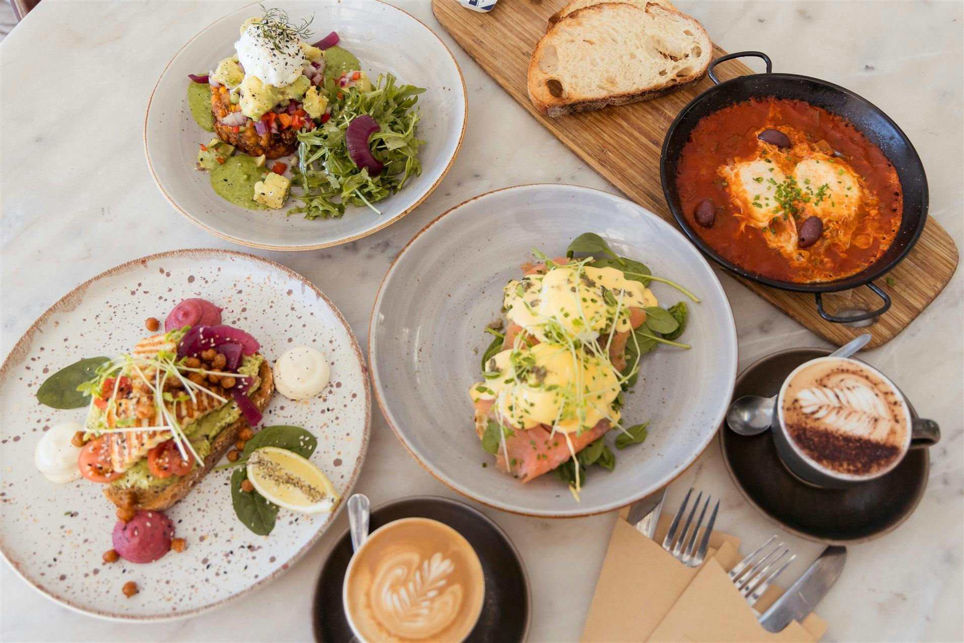 A table covered in plated eggs benedict, shakshuka, corn fritters and coffee
