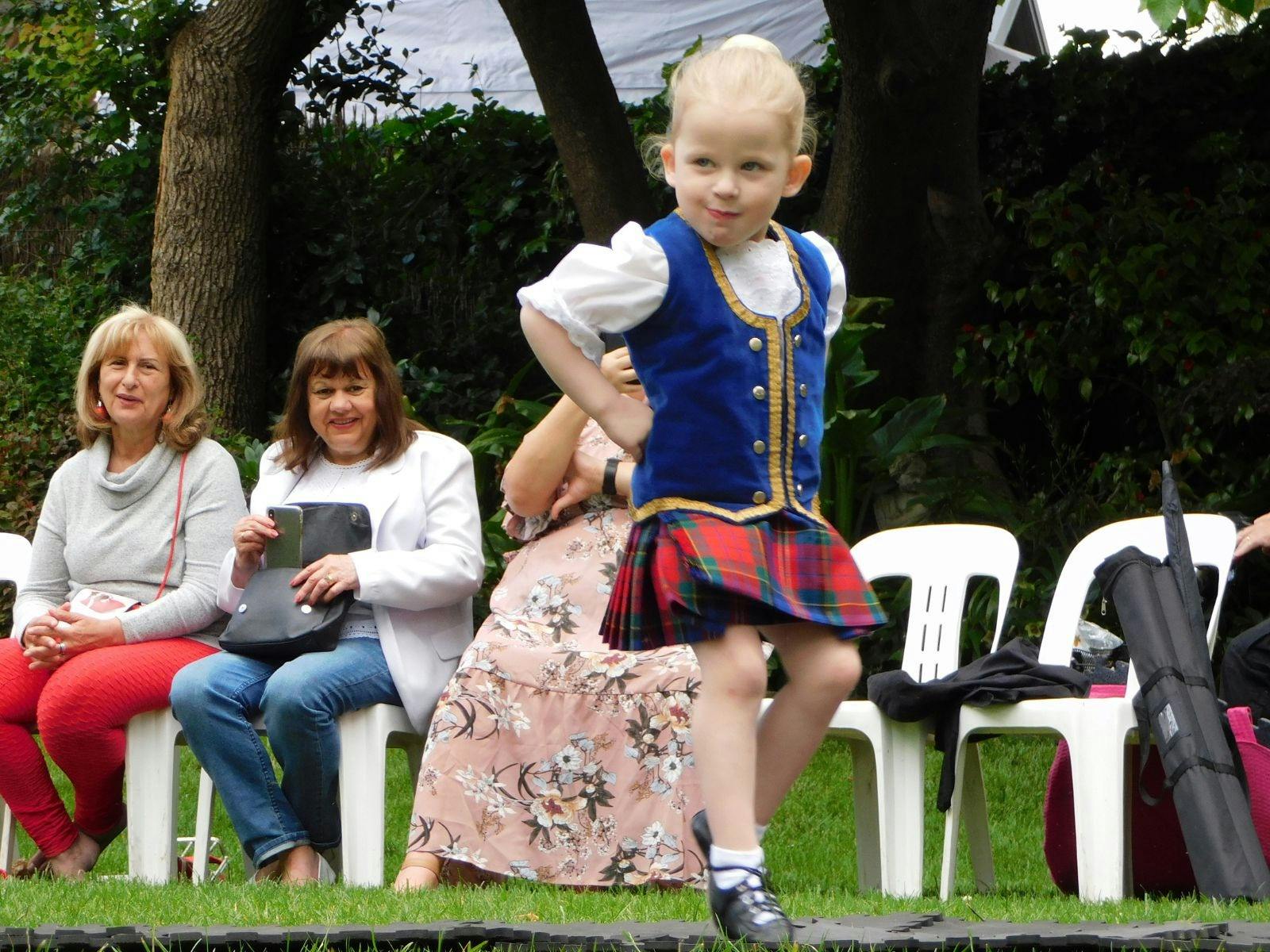 Child in Scottish traditional attire is dancing while women sitting behind her watch.